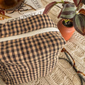 A close-up of a brown and tan checkered quilted pouch featuring a white zipper and cream lining. The pouch is styled next to a small terracotta plant and vintage-style glasses, emphasizing a cozy, handcrafted aesthetic.