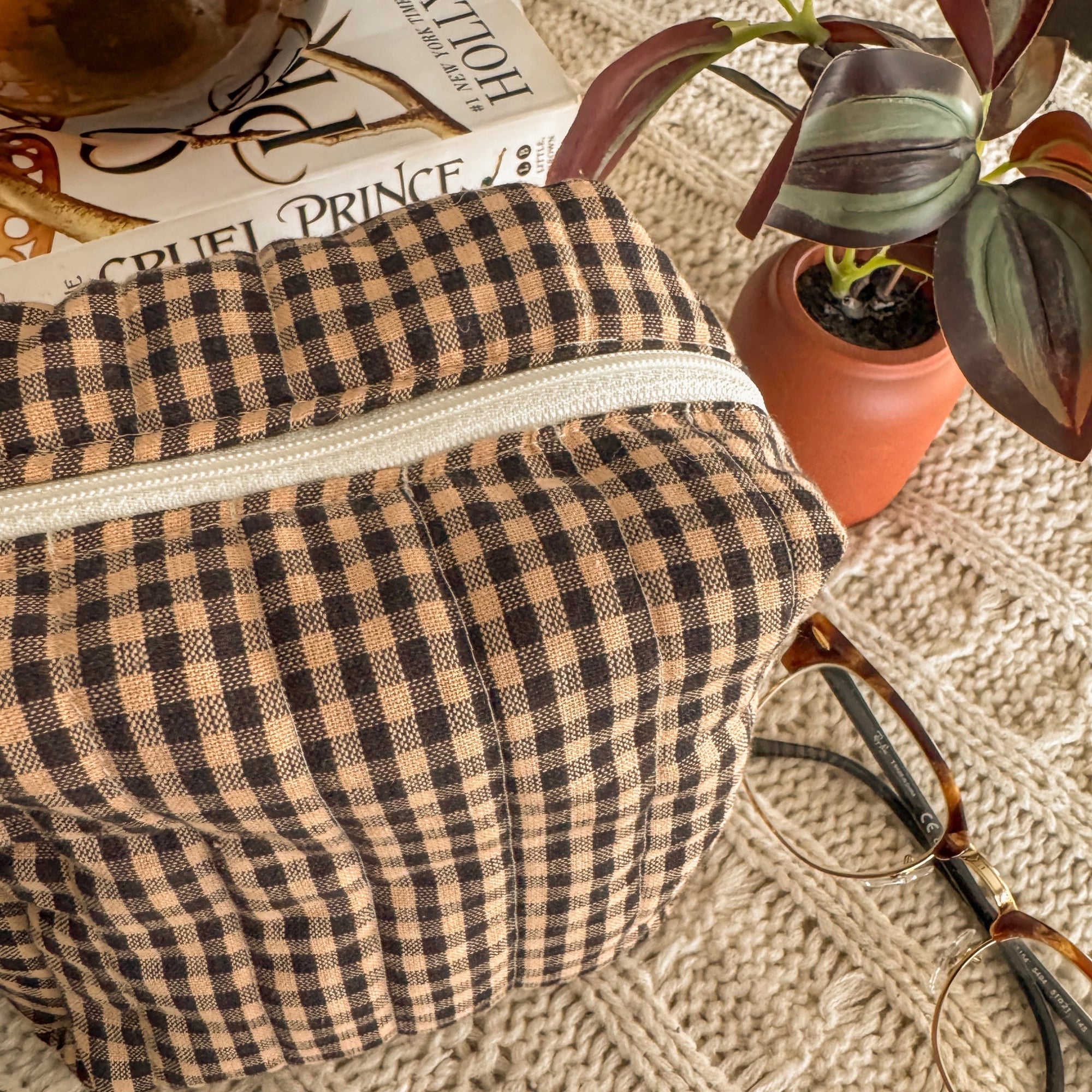 A close-up of a brown and tan checkered quilted pouch featuring a white zipper and cream lining. The pouch is styled next to a small terracotta plant and vintage-style glasses, emphasizing a cozy, handcrafted aesthetic.