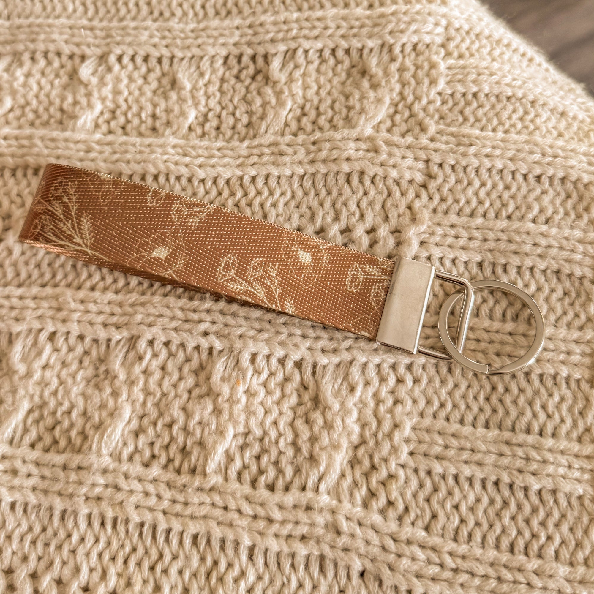 A close-up of a brown fabric key fob featuring a delicate white floral pattern and silver metal hardware. The wristlet is styled on a chunky cream knit blanket, emphasizing a cozy, autumnal aesthetic for book lovers.