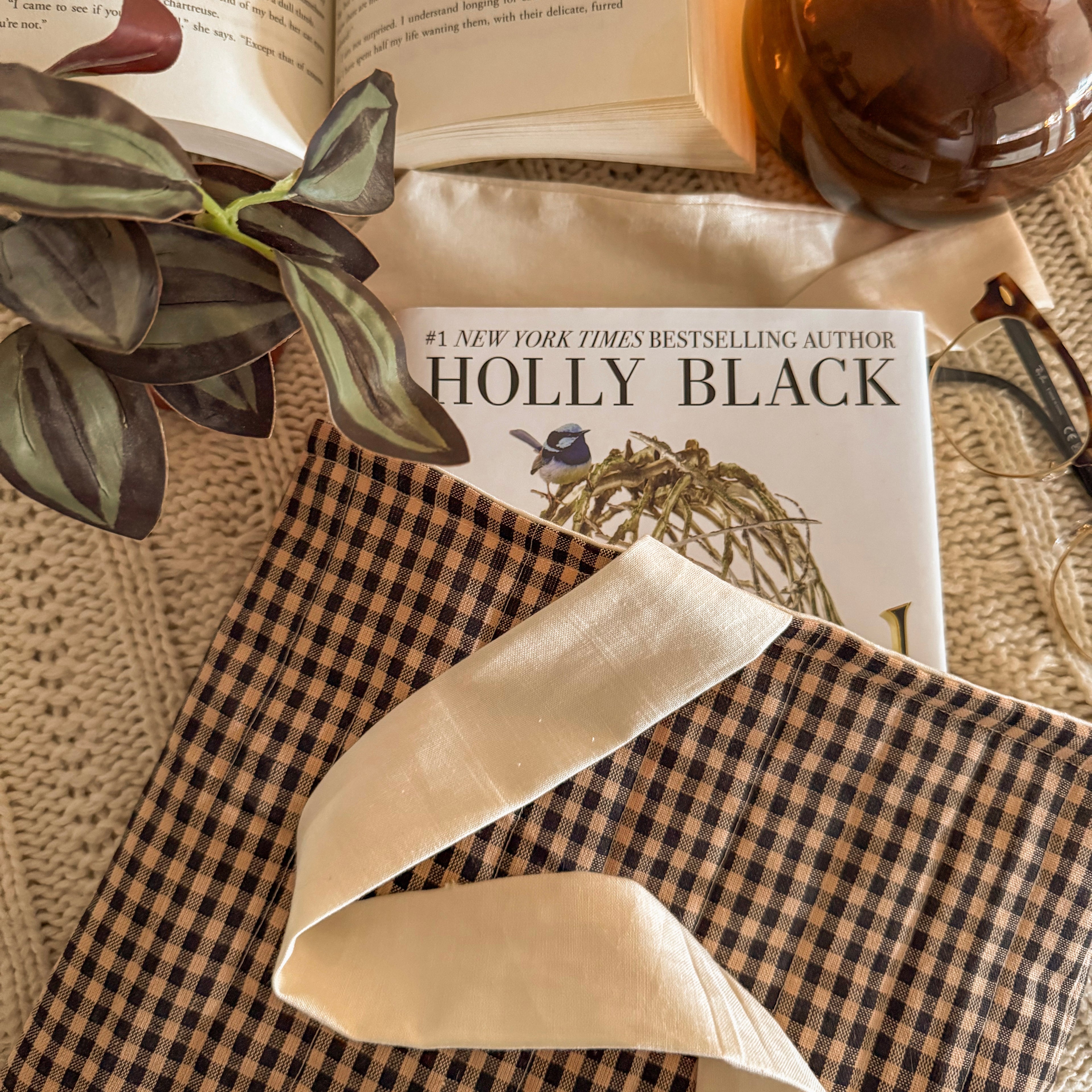 Handmade brown gingham quilted book sleeve with a cream fabric bow, protecting a Holly Black hardcover book in a cozy lifestyle setting.