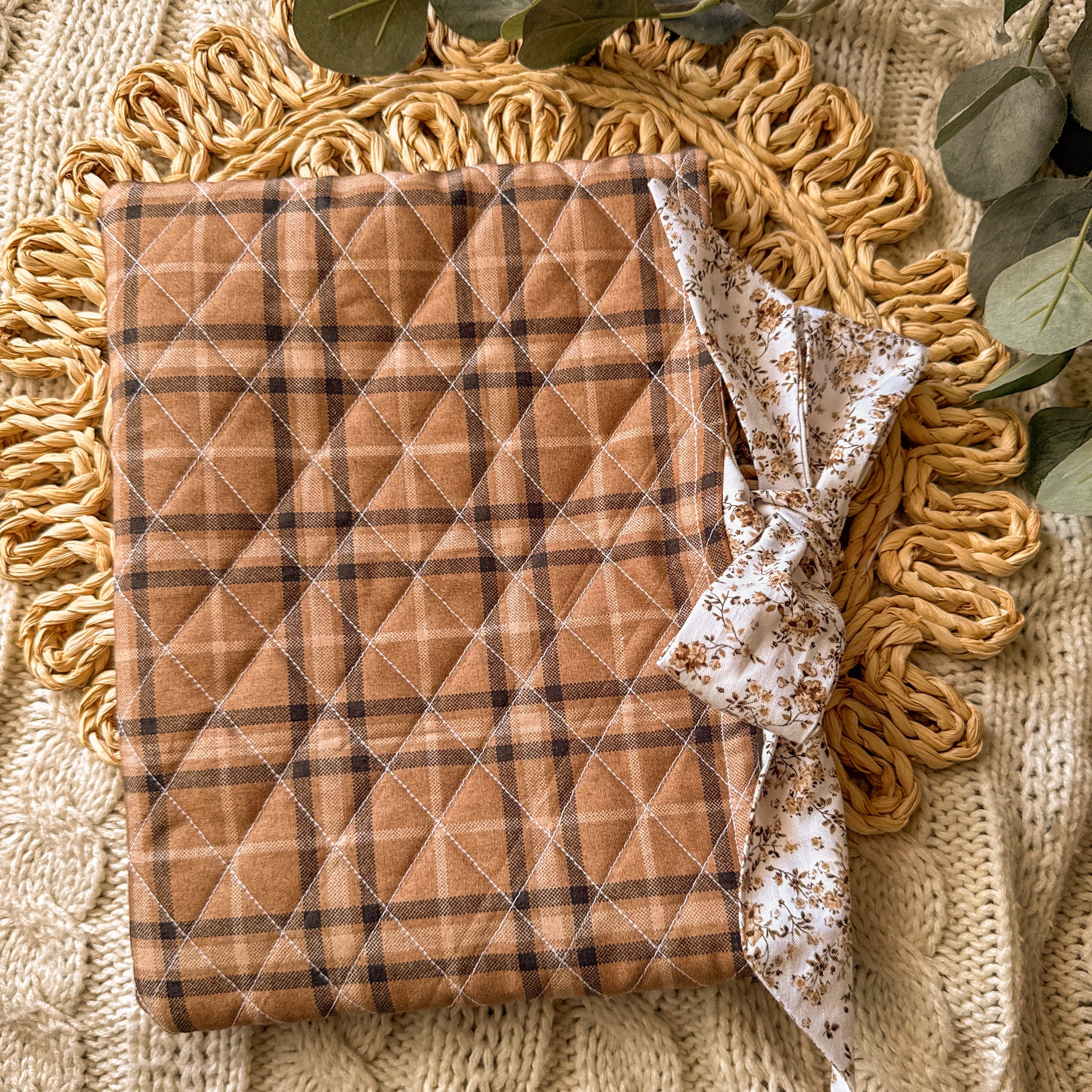 Handmade brown plaid quilted book sleeve with a decorative cream floral bow, styled on a neutral textured background with greenery for a cozy aesthetic
