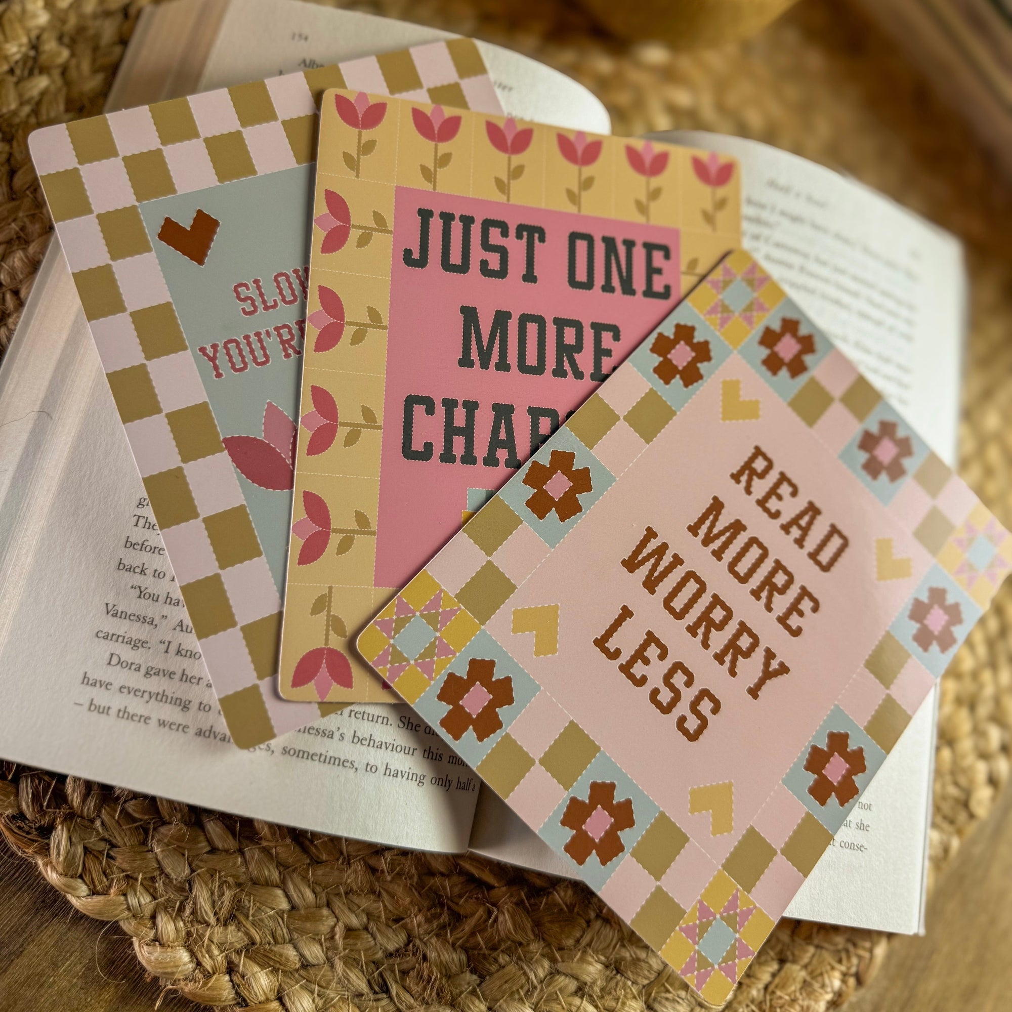 A lifestyle flat lay featuring three 'Readers' Quilt' Kindle inserts with 'Just One More Chapter,' 'Slow Down,' and 'Read More Worry Less' quotes. The inserts are fanned out on an open vintage book, styled with warm, nostalgic tones for a cozy reading aesthetic.
