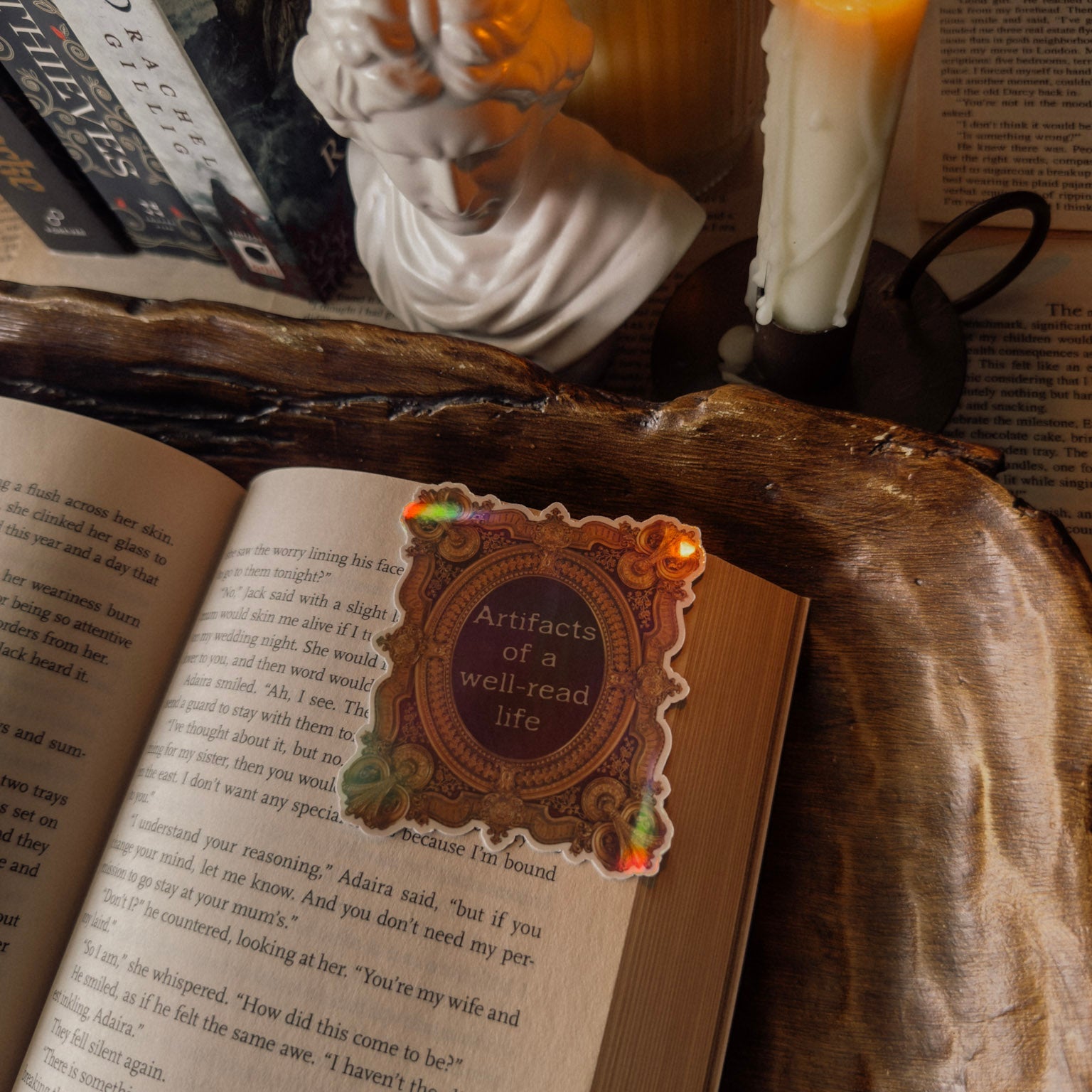Ornate holographic sticker featuring "Artifacts of a well-read life" on an open book, surrounded by dark academia decor including a candle and classical bust statue.