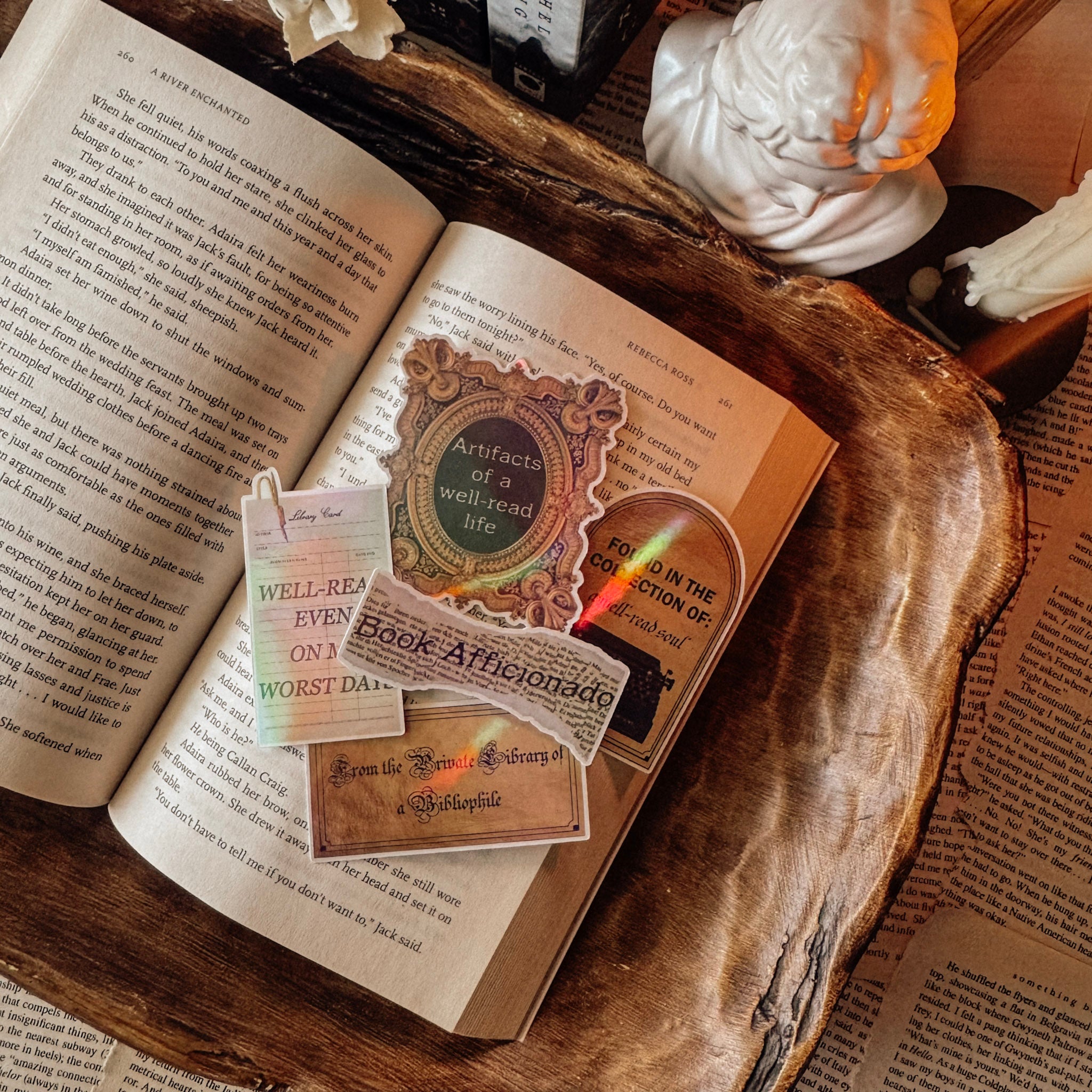 A collection of dark academia holographic stickers including "Book Aficionado," "Well-Read Soul," and "Artifacts of a Well-Read Life" designs, arranged on vintage book pages with a classical bust and candle.
