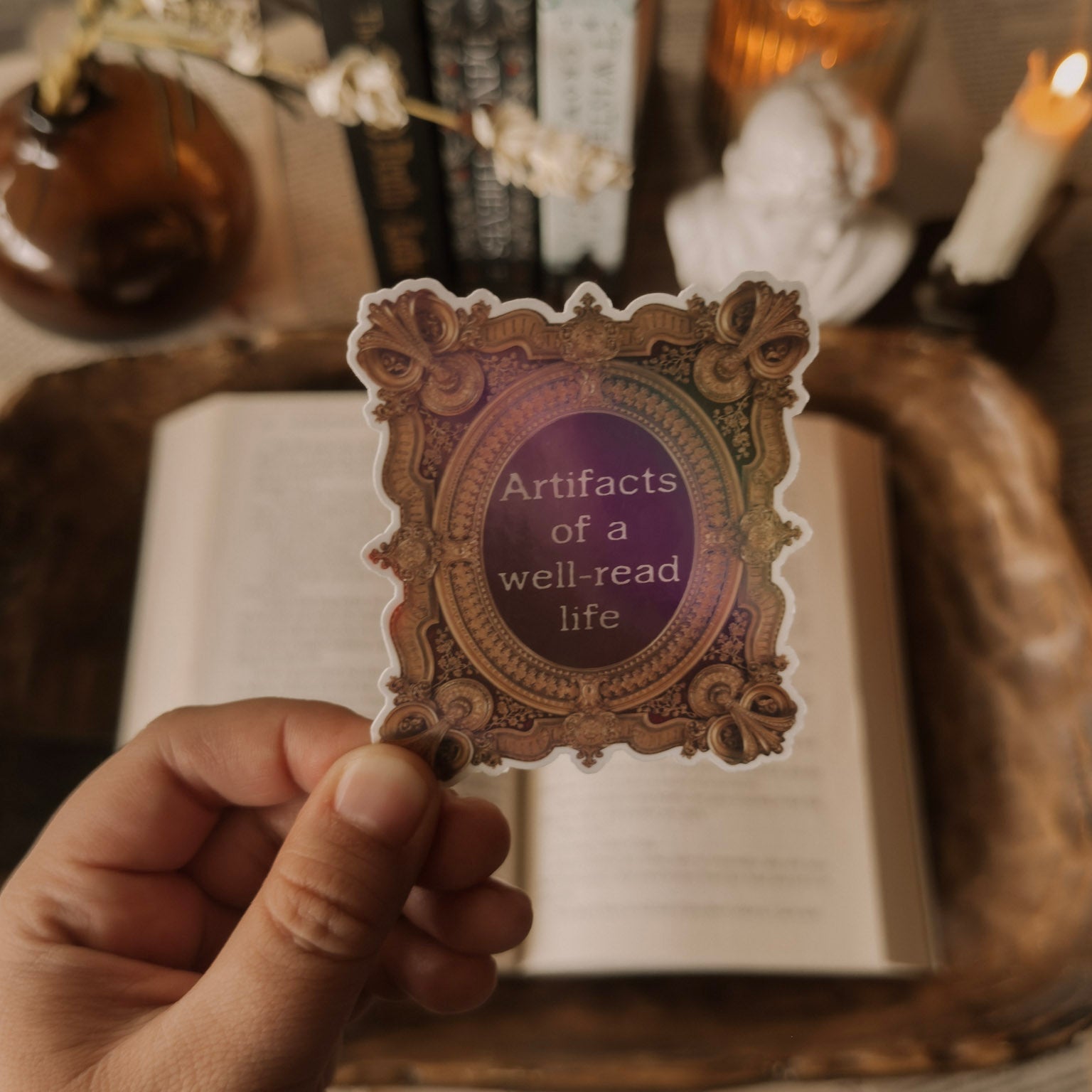 Hand holding a holographic "Artifacts of a well-read life" sticker in an ornate vintage frame against a cozy Dark Academia background with books and candles.