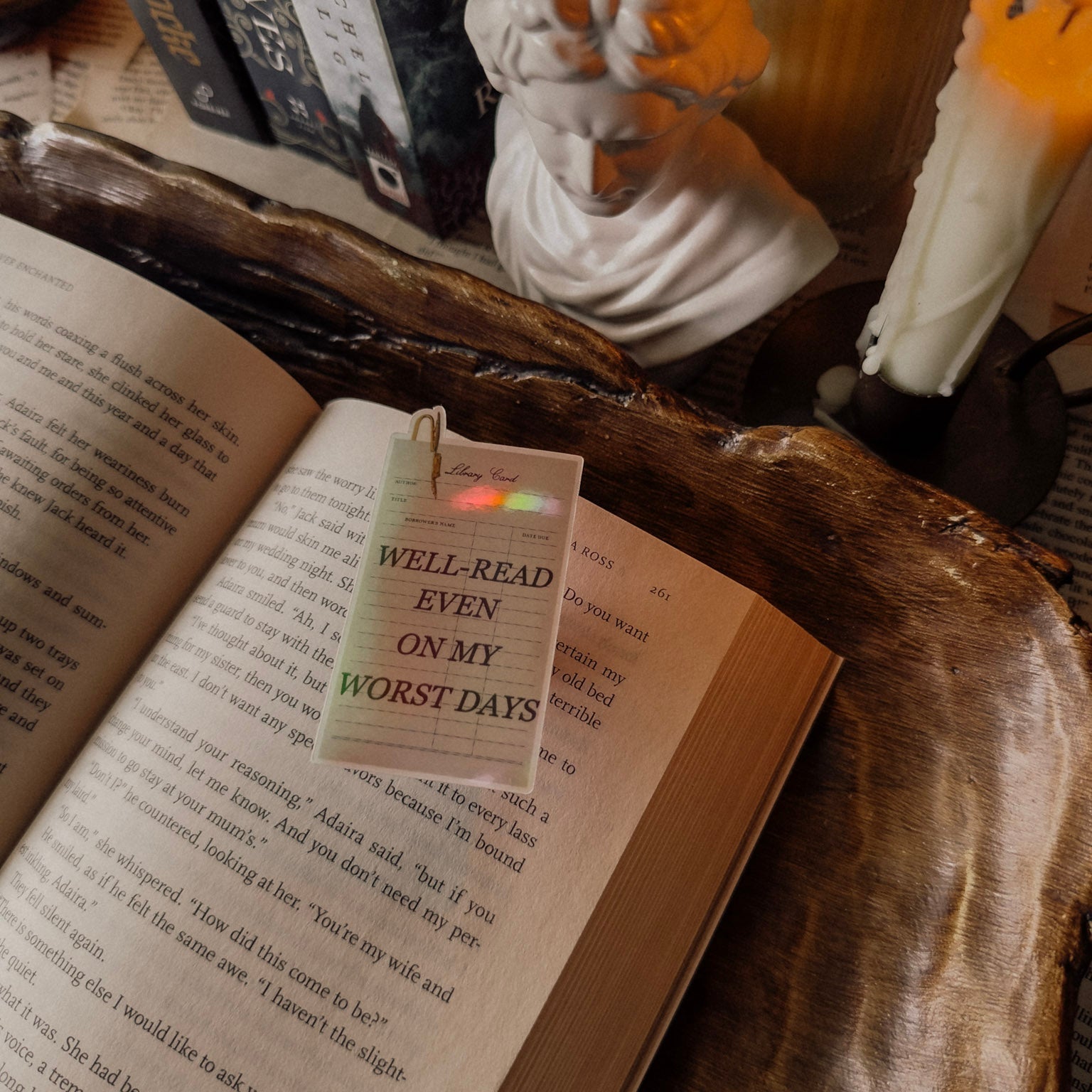 Holographic "Well-Read Even on My Worst Days" sticker placed on an open vintage book, styled with a Dark Academia aesthetic including a candle and classical statue.