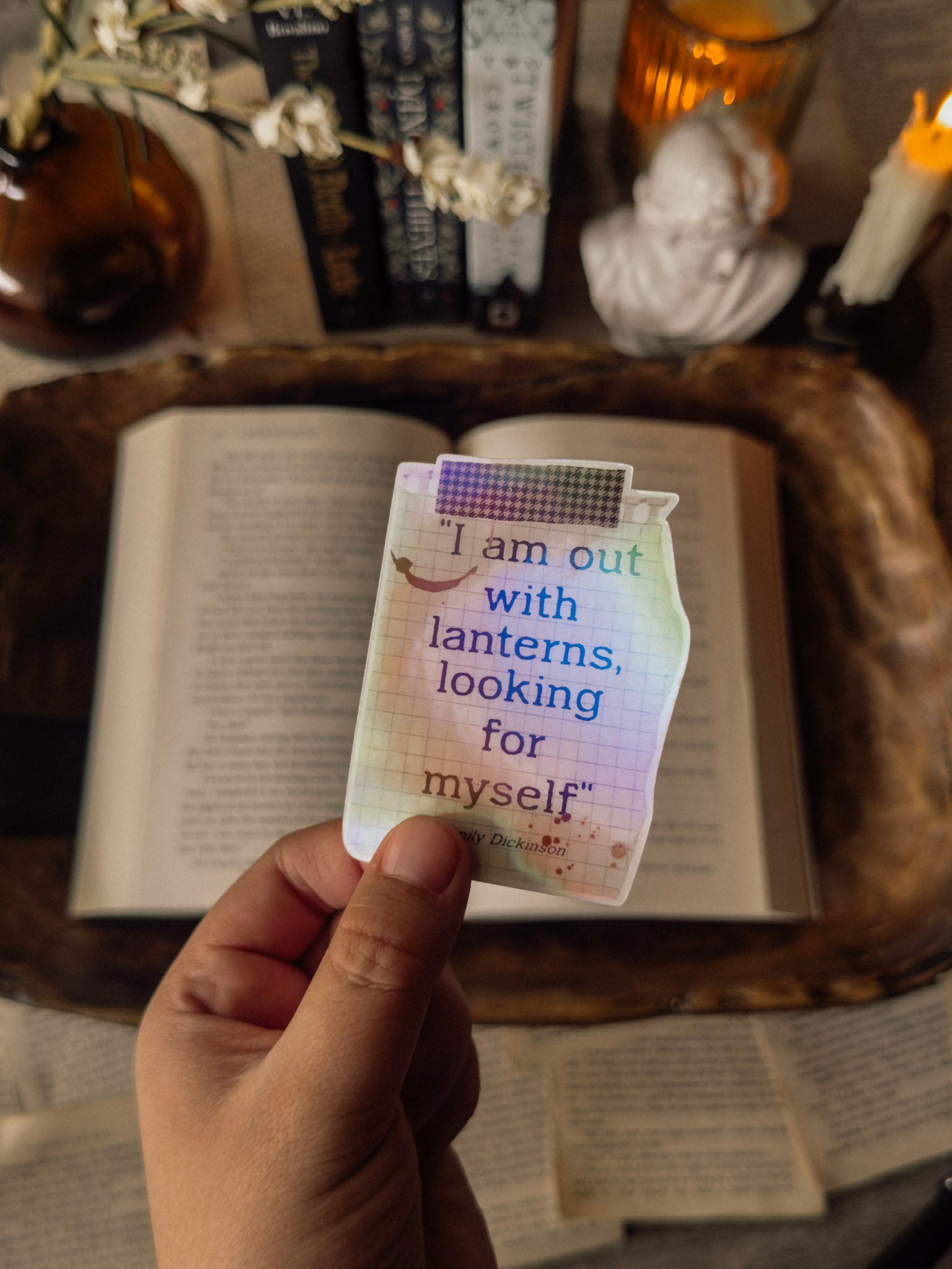 Hand holding a holographic Emily Dickinson lantern sticker to show scale. The sticker features the quote "I am out with lanterns looking for myself" and displays a vibrant rainbow holographic shimmer against a backdrop of a vintage book and marble statue.