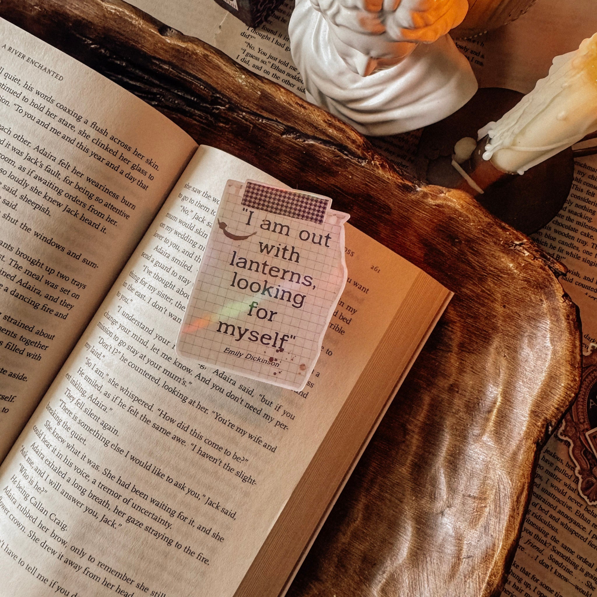 Atmospheric shot of a holographic Emily Dickinson quote sticker "I am out with lanterns looking for myself." The sticker catches a rainbow prismatic glow, styled next to a classical marble bust and a lit white candle in a moody poetcore library setting.