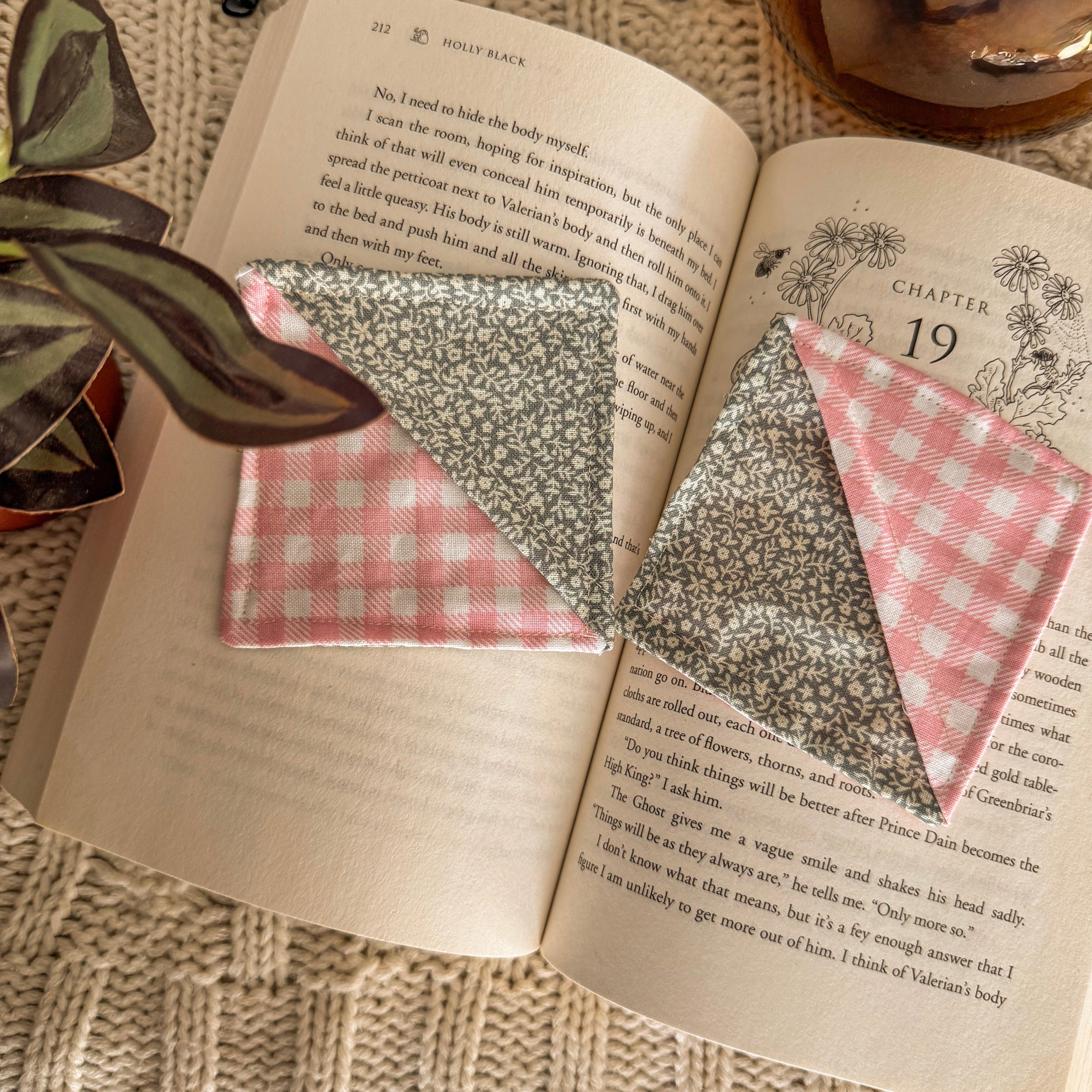 A set of two fabric corner bookmarks in green floral and pink gingham patterns, shown in use within the pages of 'The Wicked King' by Holly Black. The scene is softly lit, capturing an intentional reading ritual.