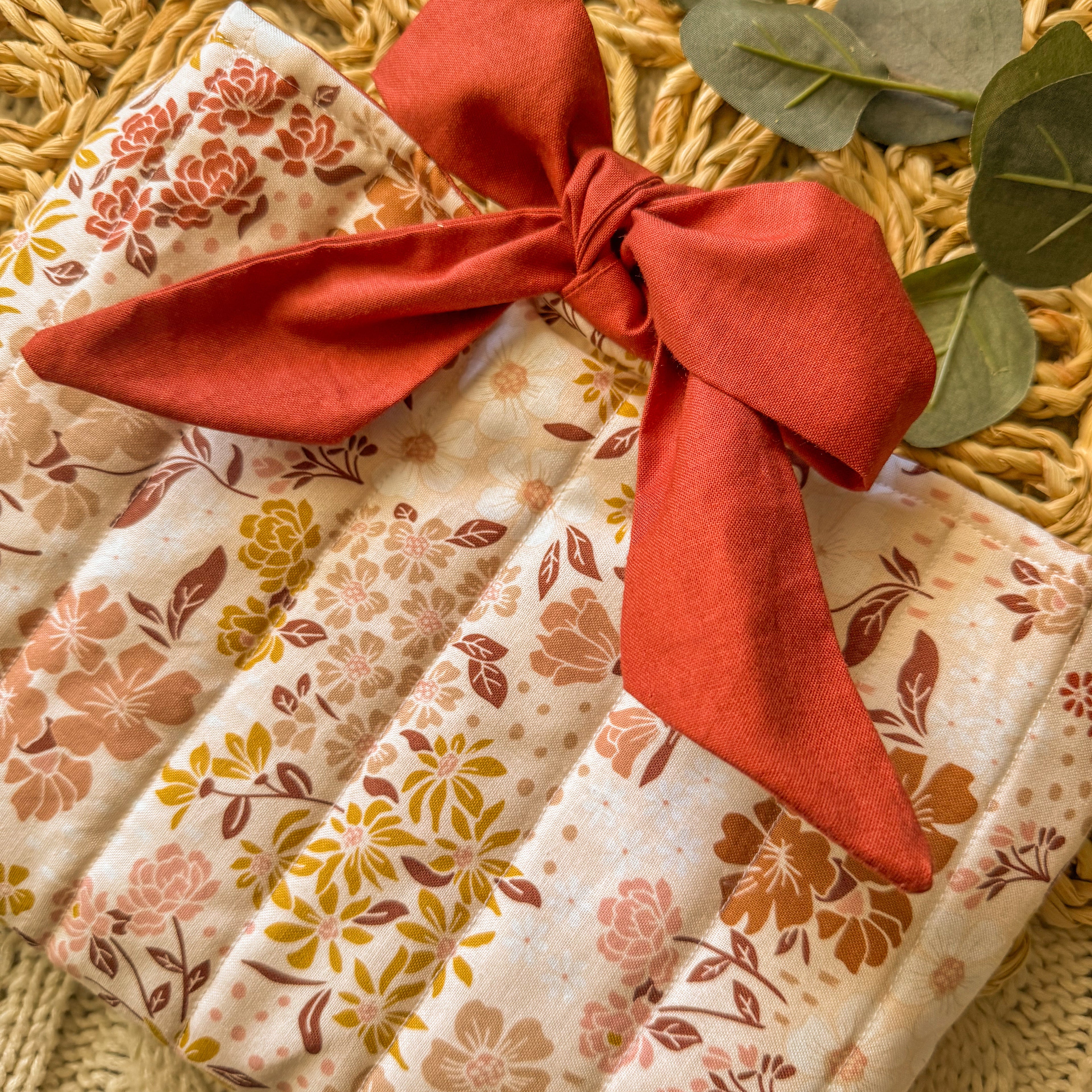 Handmade floral quilted book sleeve with a large red fabric bow, featuring a fall-inspired flower pattern on a textured background for cozy bookish protection.