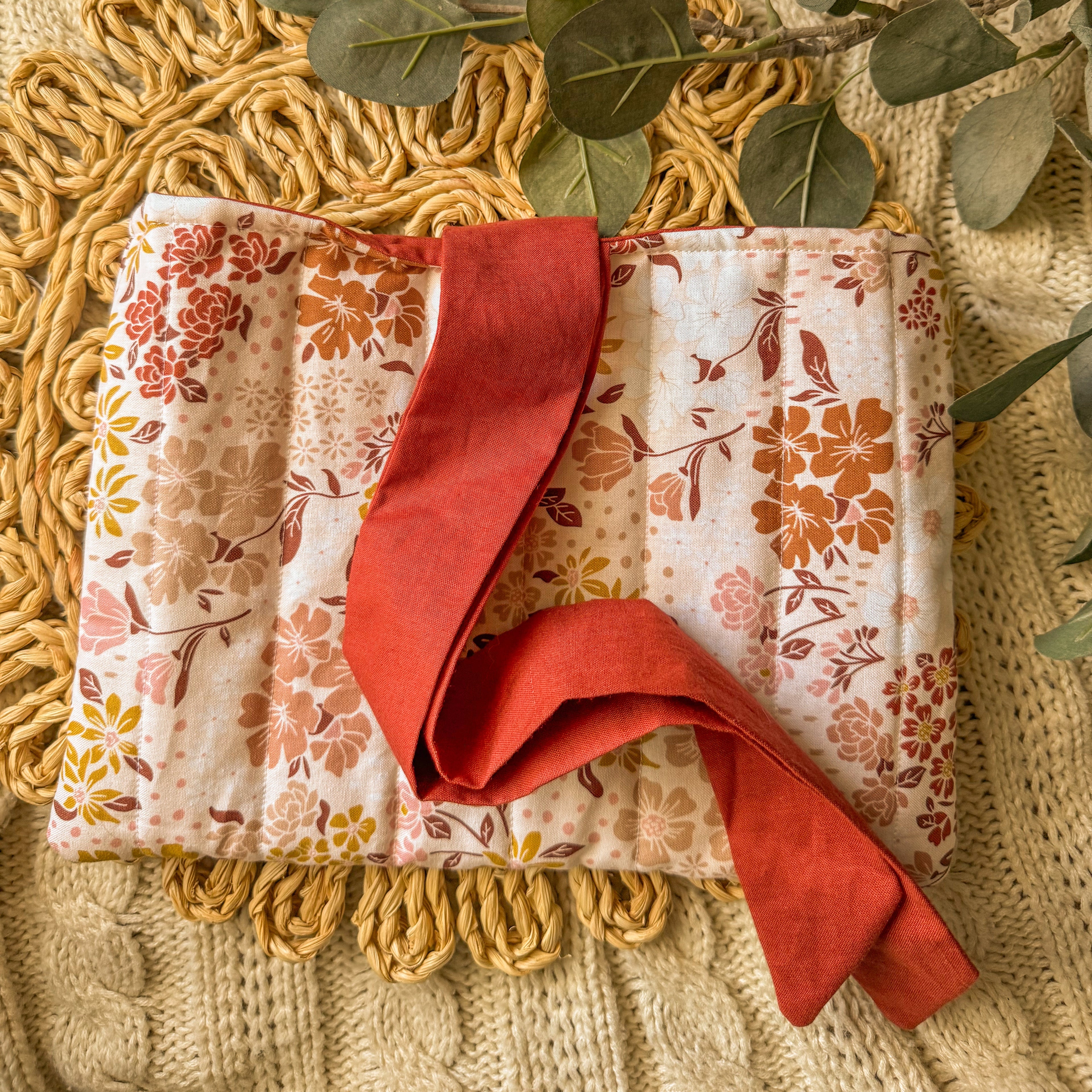 Handmade floral quilted book sleeve with a long red fabric ribbon tie, displayed on a textured cable-knit background with eucalyptus leaves for a cozy bookish aesthetic.