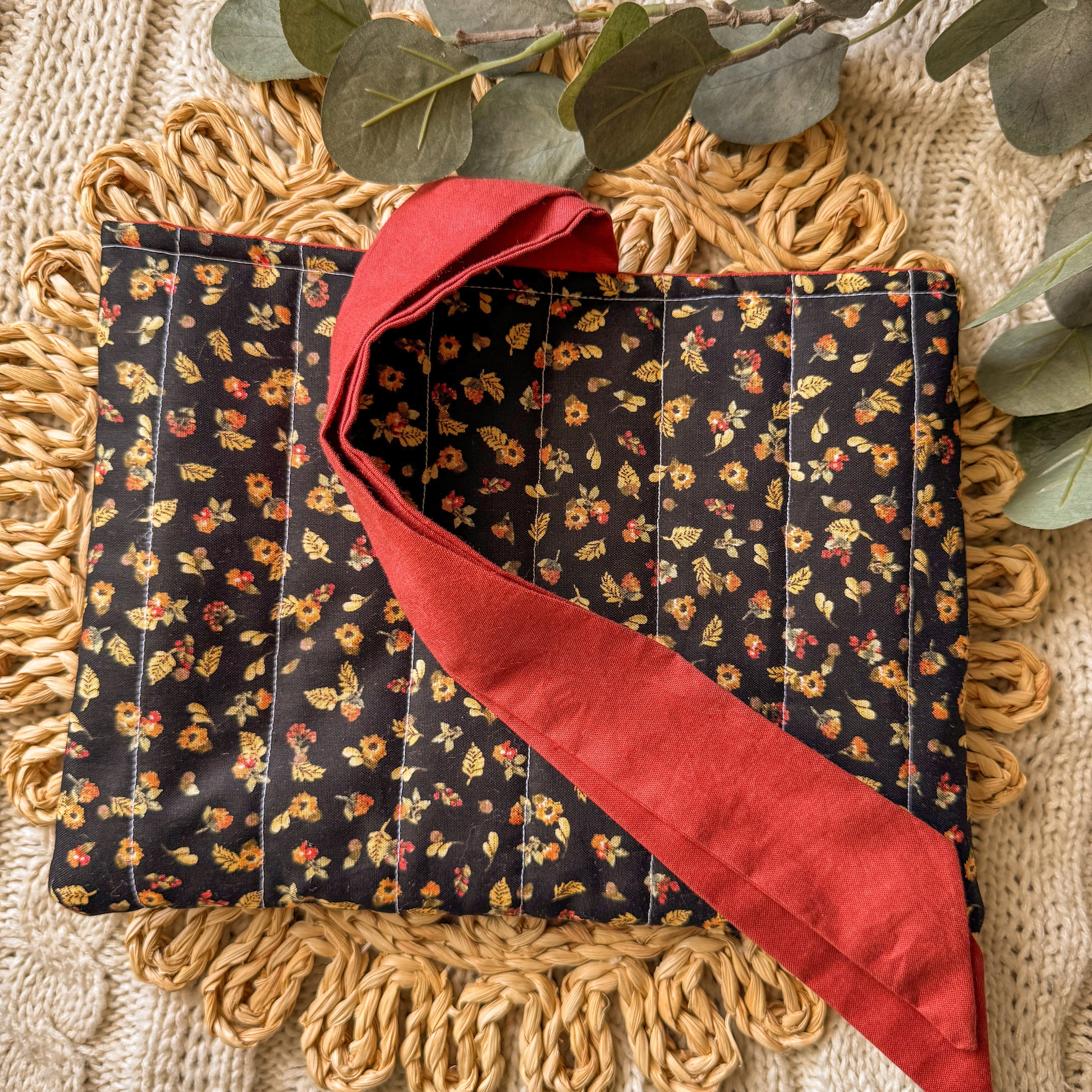 Handmade floral quilted book sleeve with a red fabric tie closure, displayed on a textured fringed mat with eucalyptus leaves for a cozy aesthetic