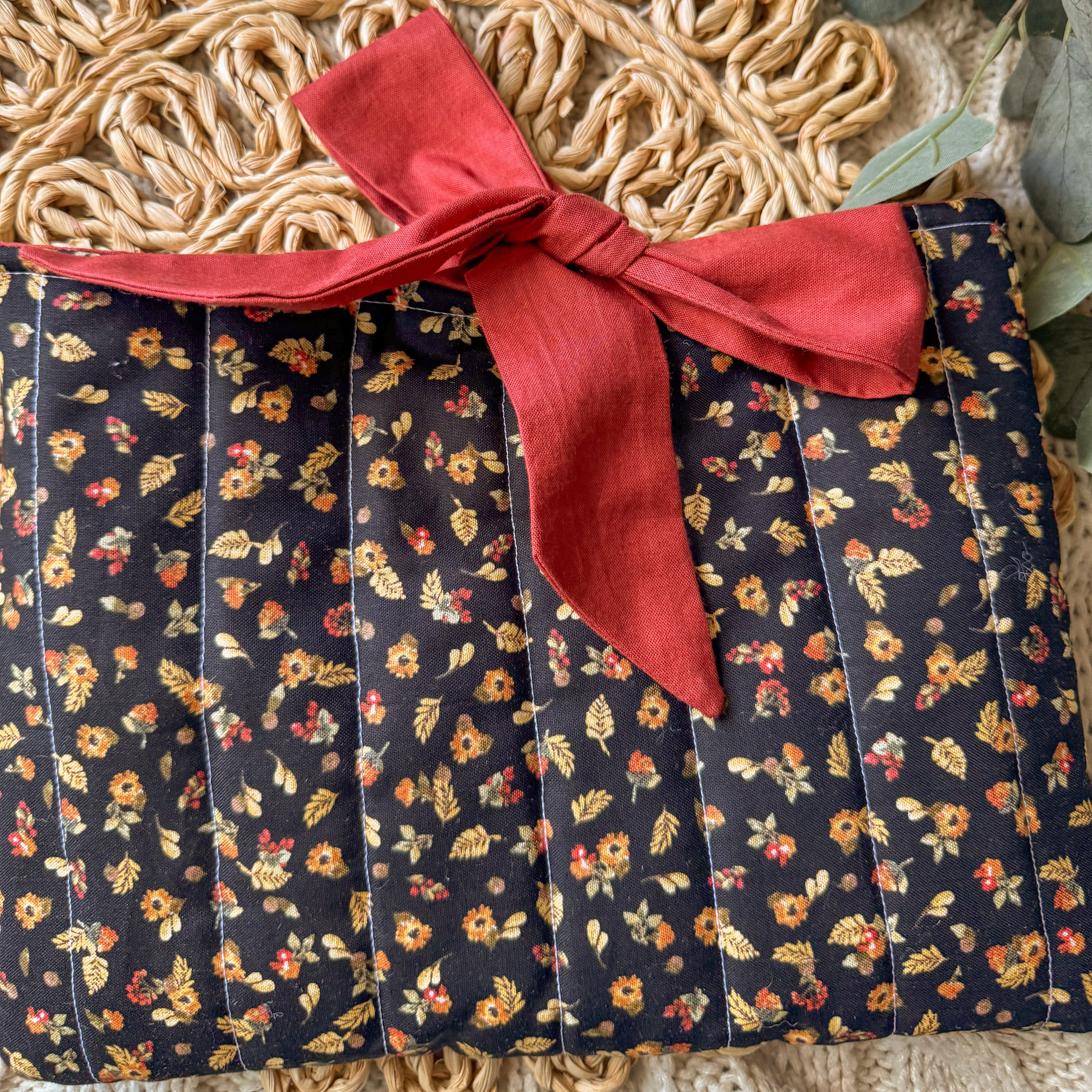 Handmade floral quilted book sleeve with a red fabric ribbon tie closure, displayed on a cozy cable-knit background with eucalyptus leaves for a bookish aesthetic