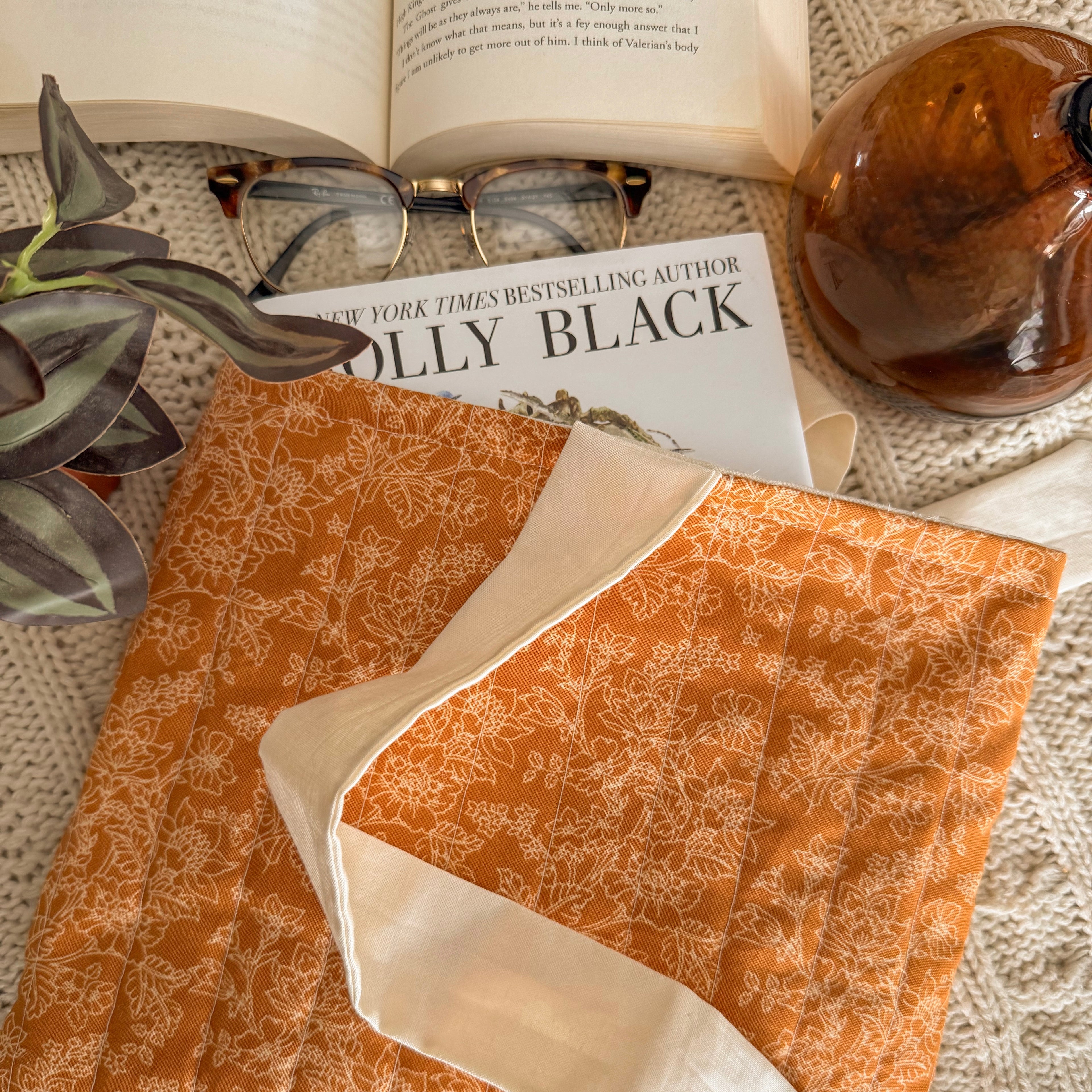 Handmade yellow and orange floral quilted book sleeve protecting a Holly Black hardcover book, styled in a cozy flat lay with glasses and a plant