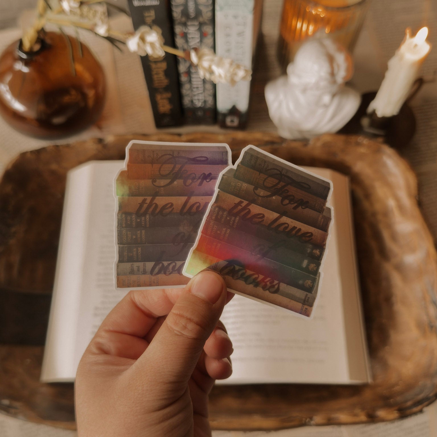 Holographic "For the Love of Books" sticker featuring a stack of vintage books. Styled in a light academia setting with a white marble statue, a lit candle, and a warm prismatic rainbow shimmer.