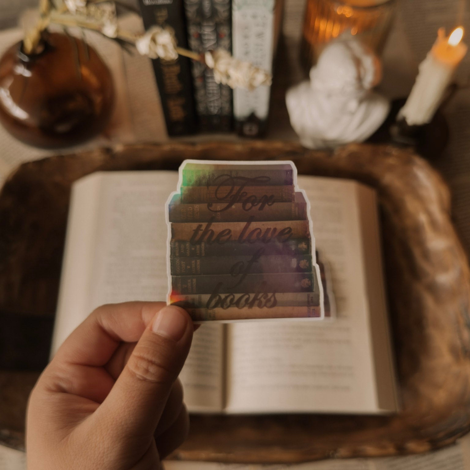 Holographic "For the Love of Books" sticker with a prismatic rainbow finish, featuring a stack of vintage novels. Styled on an open book next to a white marble Greek bust and a lit candle in a sunlit light academia setting.