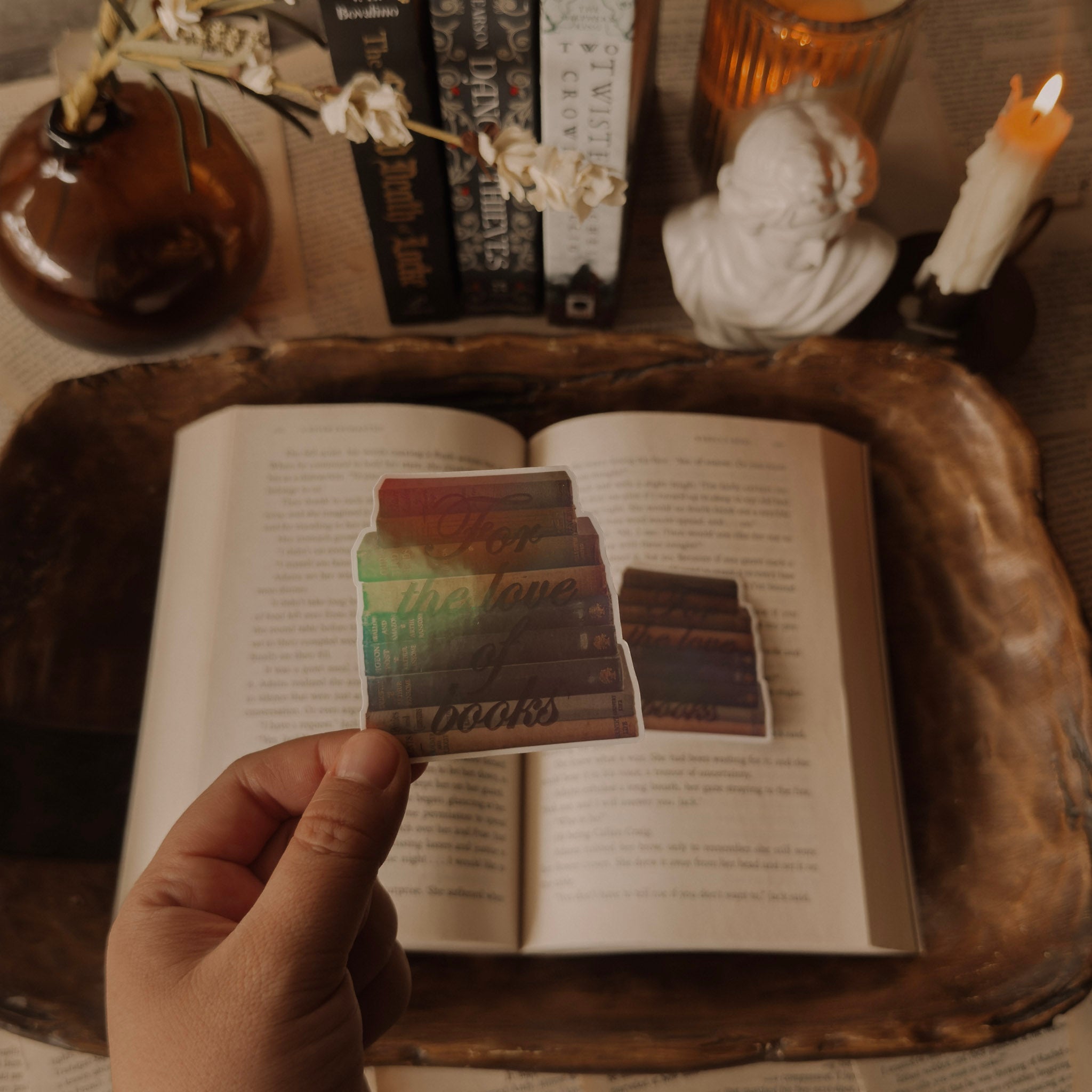 A holographic sticker titled "For the Love of Books" featuring an illustration of a stack of vintage books. The sticker is styled in a light academia setting on a weathered book page with a classical marble bust and a lit candle in the background.
