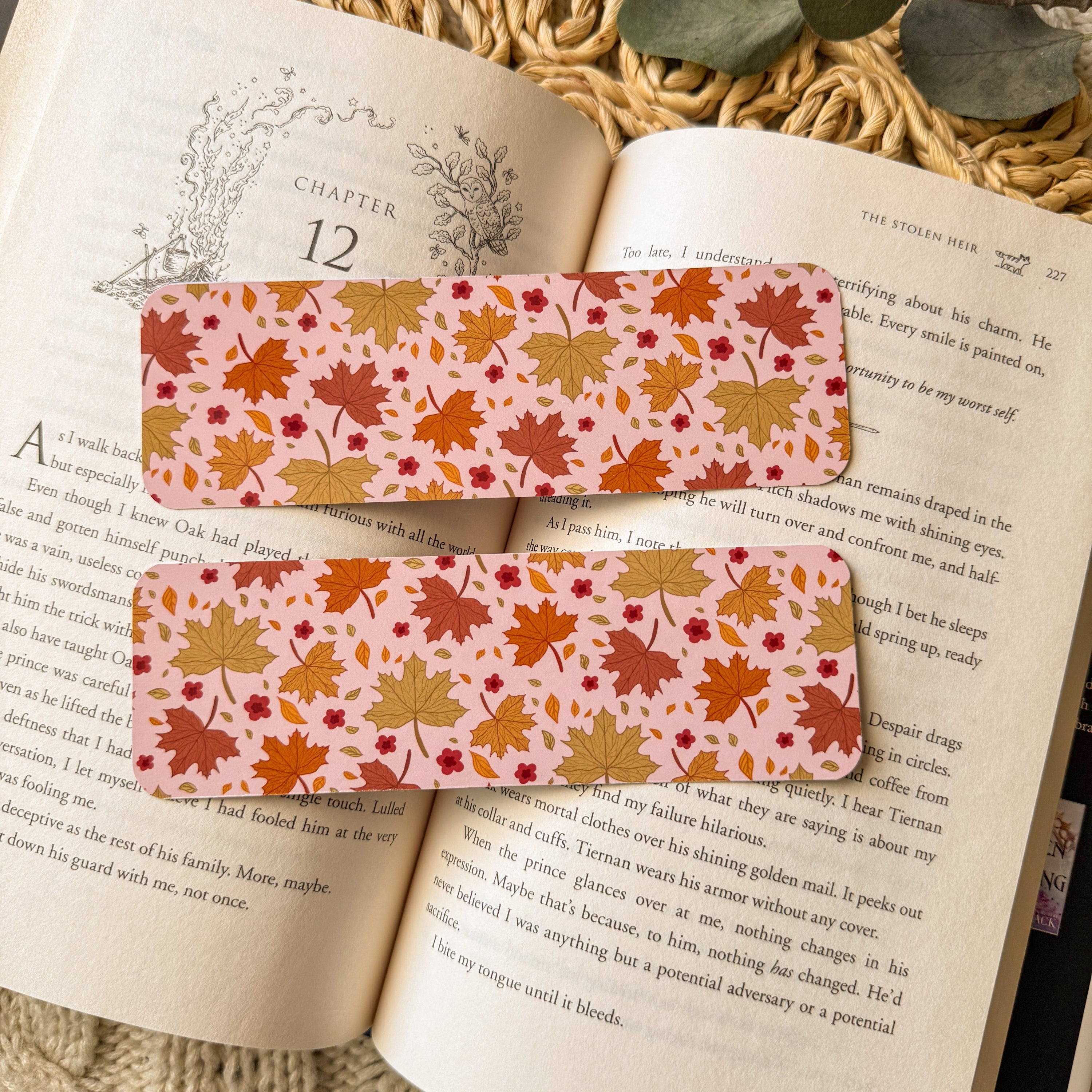 Two glossy bookmarks with a warm golden botanical pattern resting on the pages of an open book. The lifestyle shot captures a quiet reading moment with natural textures and soft light.