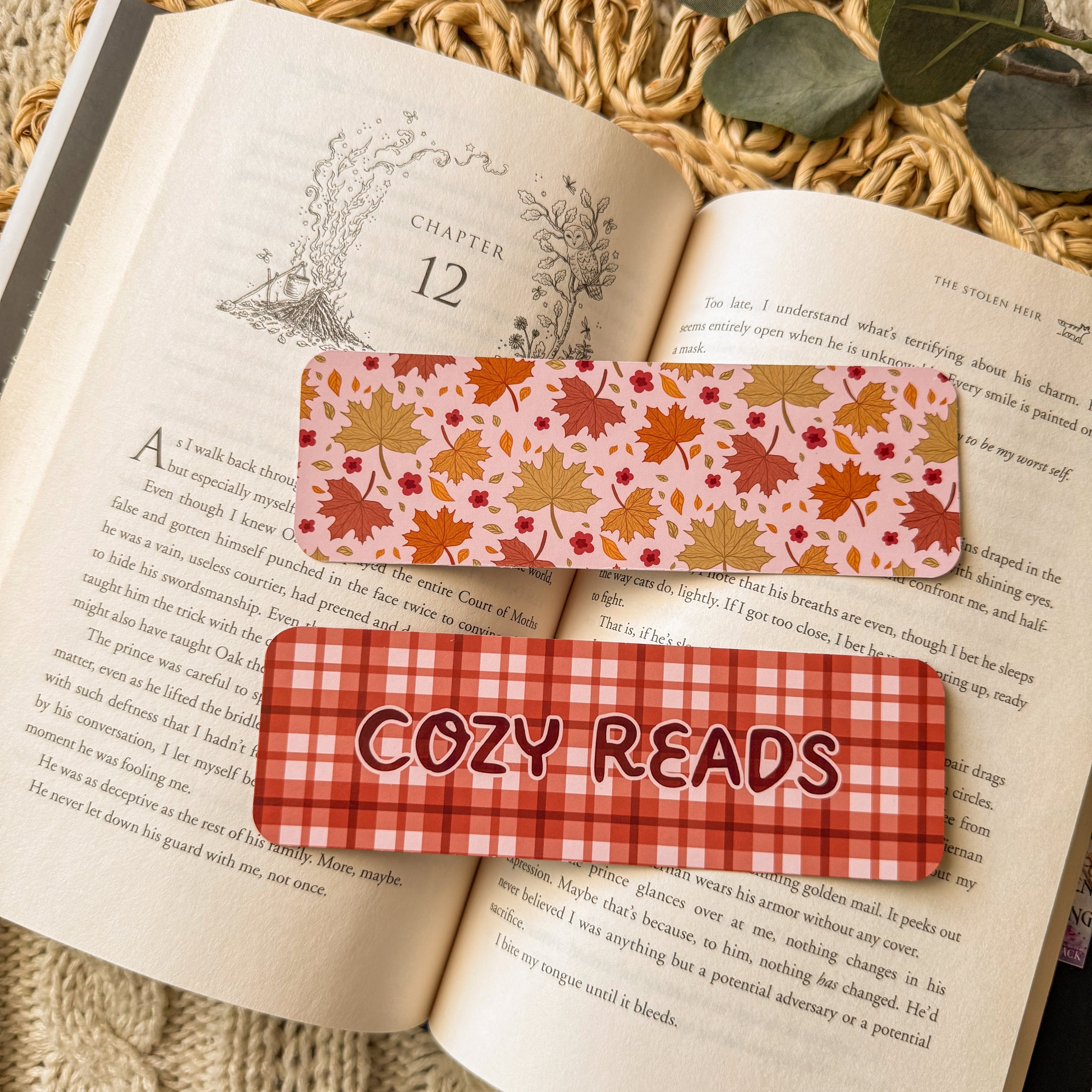 Two glossy bookmarks featuring warm, light-filled patterns and the text 'Cozy Reads,' resting on the pages of an open book with a textured background