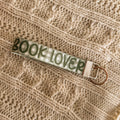 A close-up of a green gingham fabric key fob featuring the text 'BOOK LOVER' in dark green lettering. The wristlet is styled on a cream-colored cable knit sweater, highlighting its soft texture and cozy literary aesthetic.