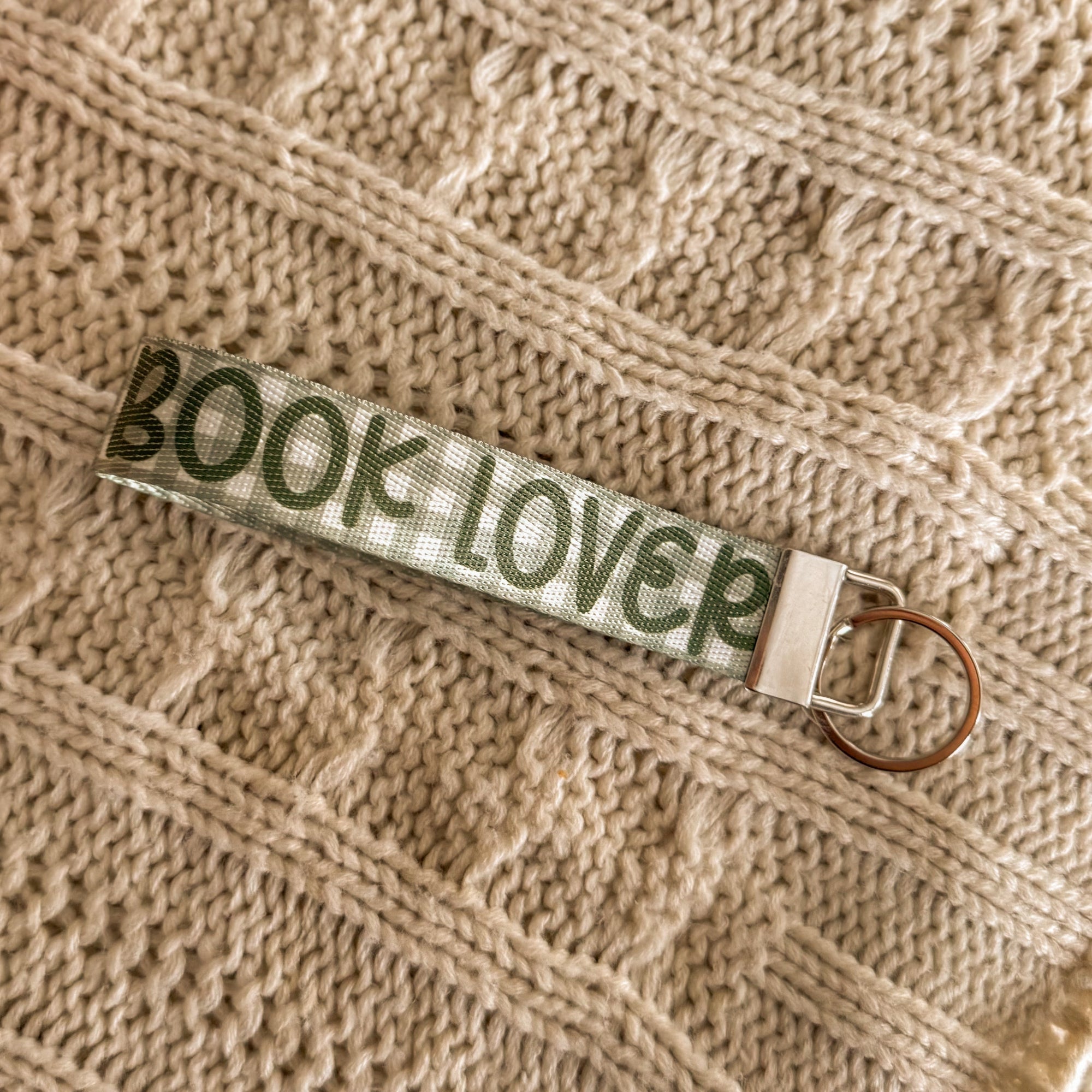 A close-up of a green gingham fabric key fob featuring the text 'BOOK LOVER' in dark green lettering. The wristlet is styled on a cream-colored cable knit sweater, highlighting its soft texture and cozy literary aesthetic.
