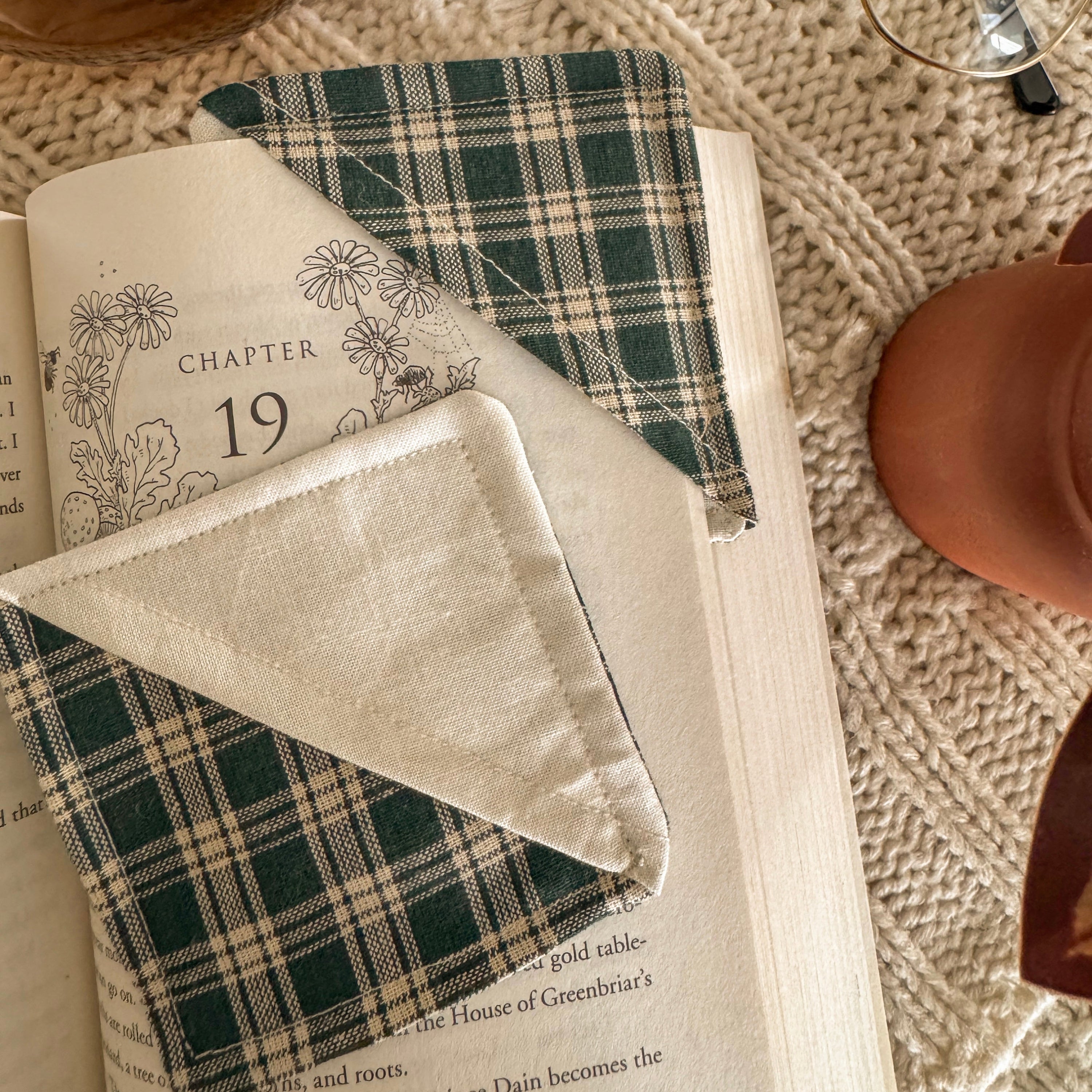 A close-up of a green plaid fabric corner bookmark holding a place in a book at Chapter 19. The dark green and cream pattern is shown against the off-white pages of an open book in a dimly lit, archival setting