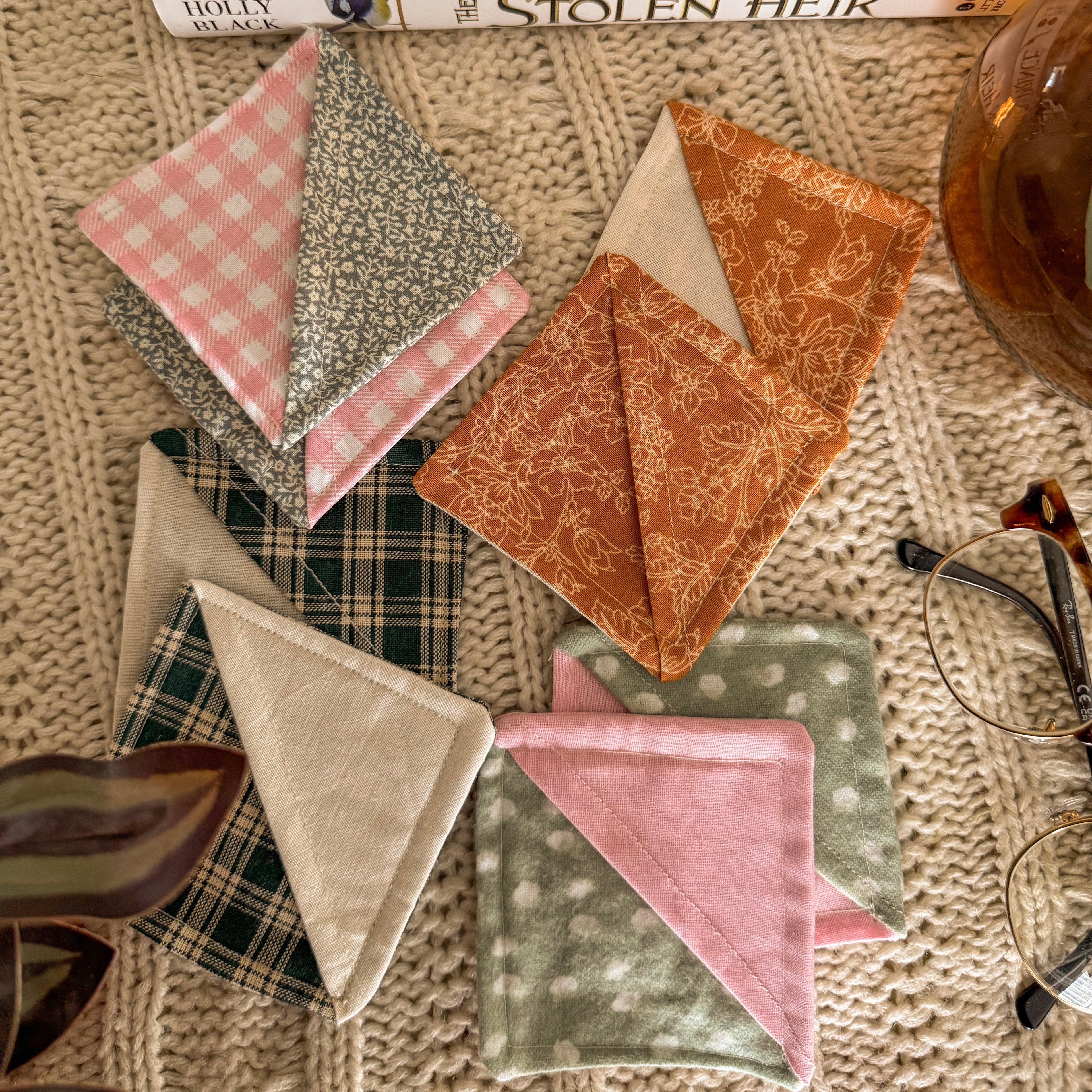 A flat lay featuring four pairs of handmade fabric corner bookmarks in various floral, plaid, and gingham patterns. Styled on a cream knit blanket with a book and vintage-style glasses for an archival aesthetic