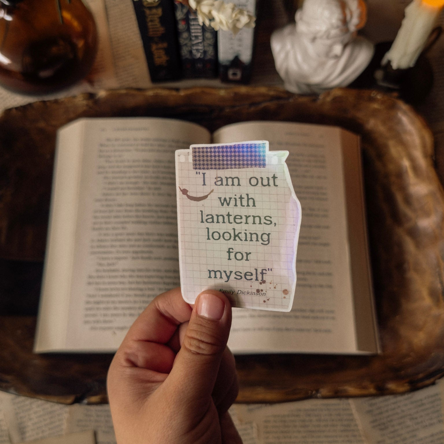 Holographic literary sticker with a vintage lantern illustration and the Emily Dickinson quote "I am out with lanterns looking for myself." Photographed in a dark academia aesthetic on an antique book page with a marble bust and a prismatic rainbow glow.