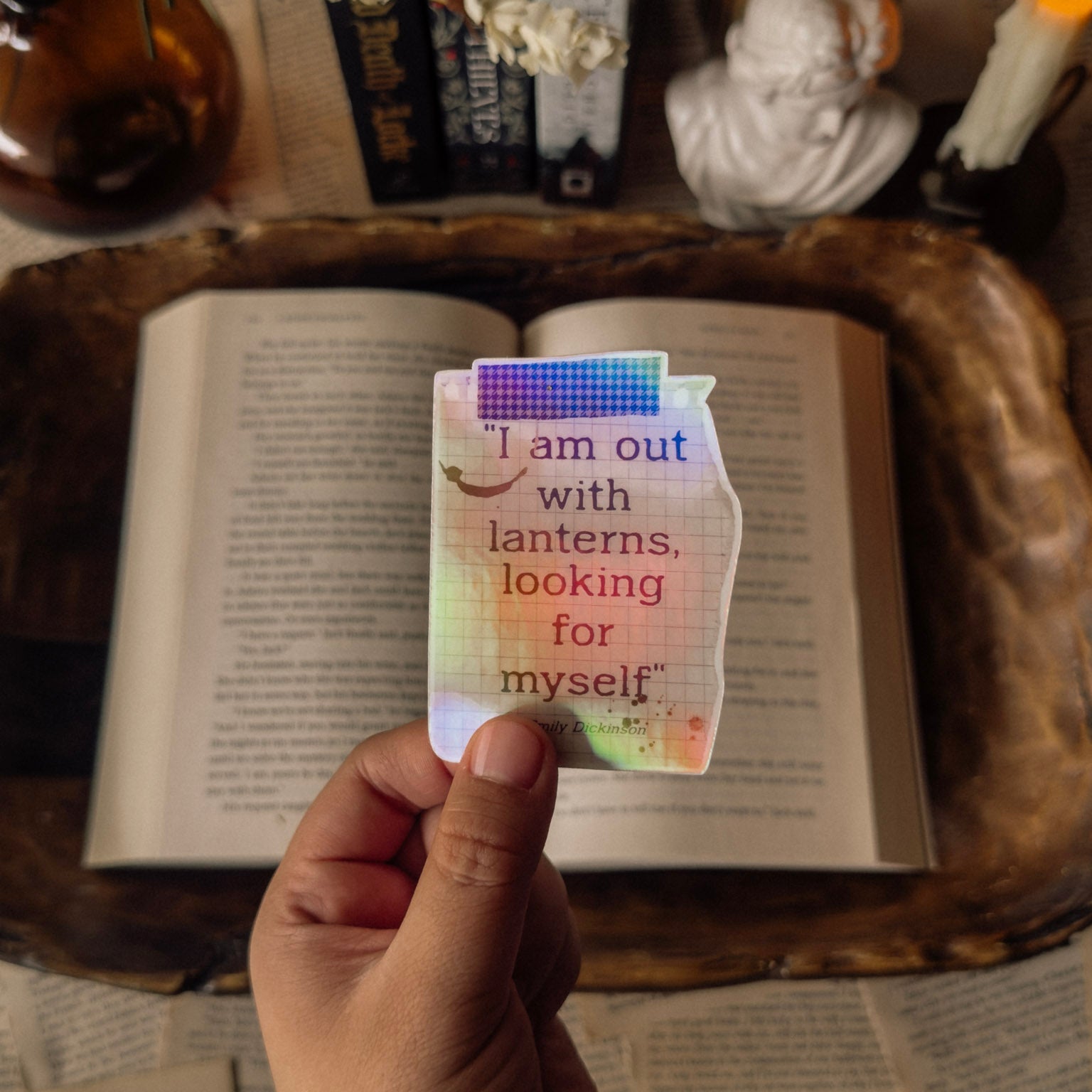 Holographic literary sticker featuring the Emily Dickinson quote "I am out with lanterns looking for myself." Illustrated with a vintage scrapbook paper in a dark academia aesthetic, styled on a weathered book with a marble bust and flickering candle.