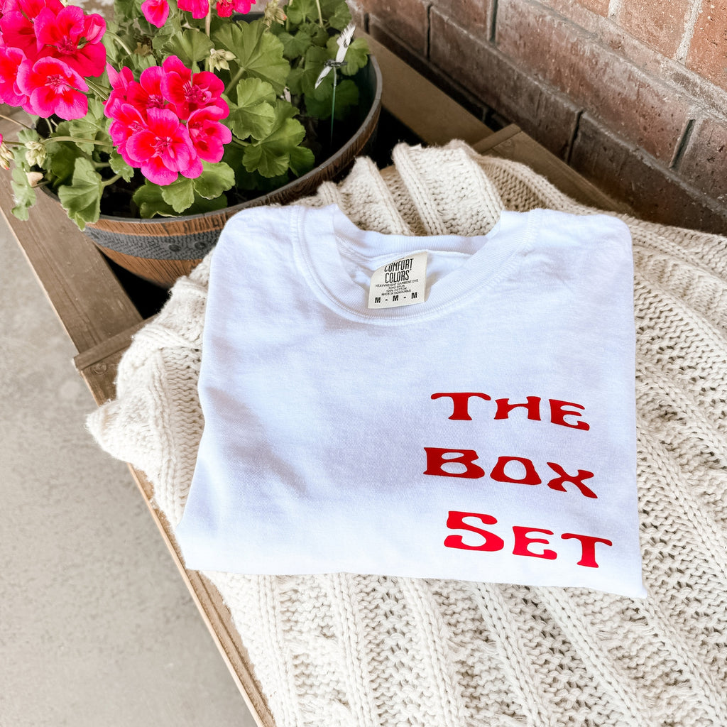 White “The Box Set” T-shirt with coupe glass stack and bold red lettering laid flat on a cozy knit blanket next to a pink flower pot on a wood bench.