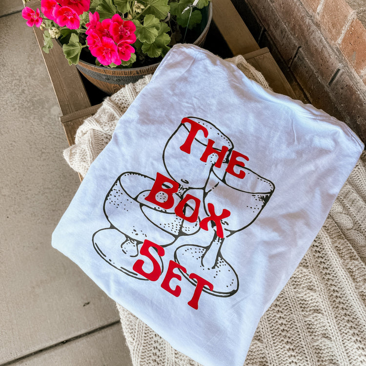 White T-shirt with red “The Box Set” text over a stacked coupe glass illustration, styled with pink flowers and cozy textures.