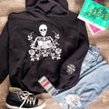Black hoodie with a central skeleton design holding a book, surrounded by floral patterns. The hoodie is displayed alongside a pair of blue jeans, a pair of black sneakers, and a few colorful books.