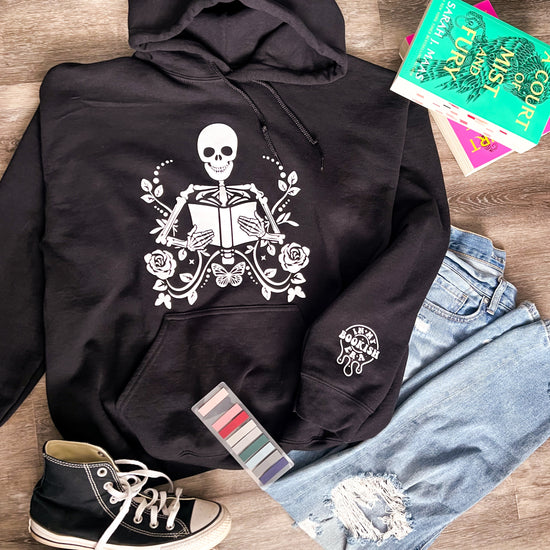 Black hoodie with a central skeleton design holding a book, surrounded by floral patterns. The hoodie is displayed alongside a pair of blue jeans, a pair of black sneakers, and a few colorful books.