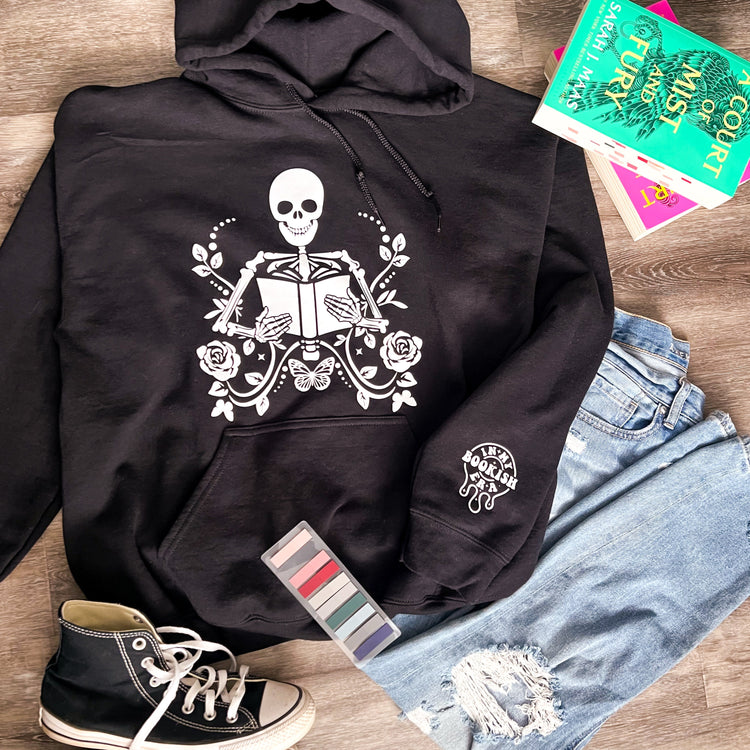 Black hoodie with a central skeleton design holding a book, surrounded by floral patterns. The hoodie is displayed alongside a pair of blue jeans, a pair of black sneakers, and a few colorful books.