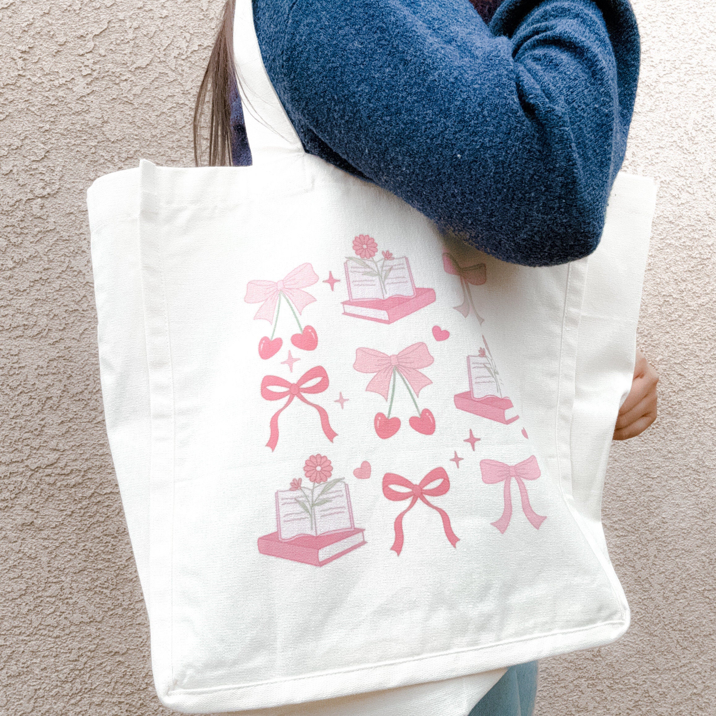 A pink canvas tote bag with a whimsical design of bows, cherries, and library-themed items like books and hearts, being held by a person with a blue sweater.