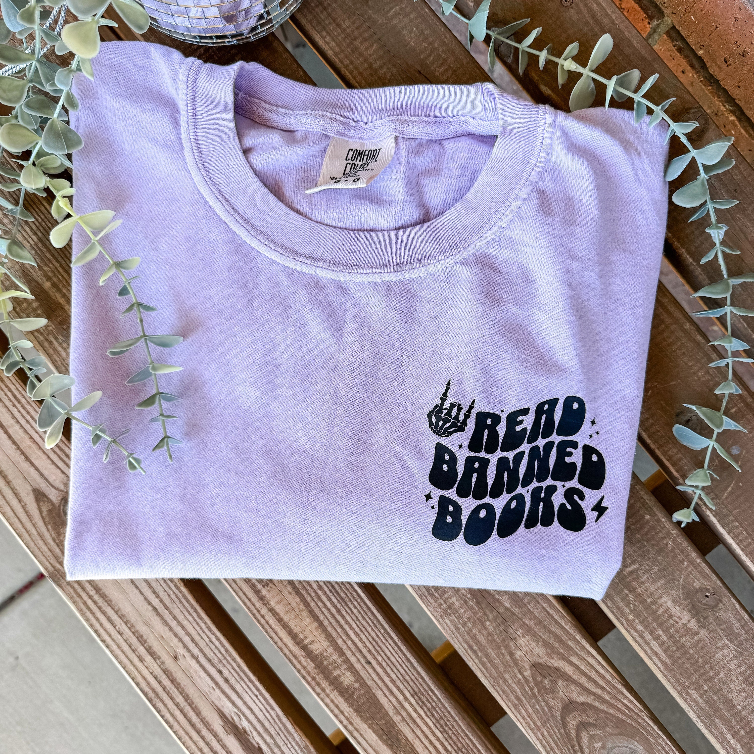 A lavender-colored T-shirt with the text 'Read Banned Books' printed on the front pocket, displayed on a wooden surface surrounded by foliage.