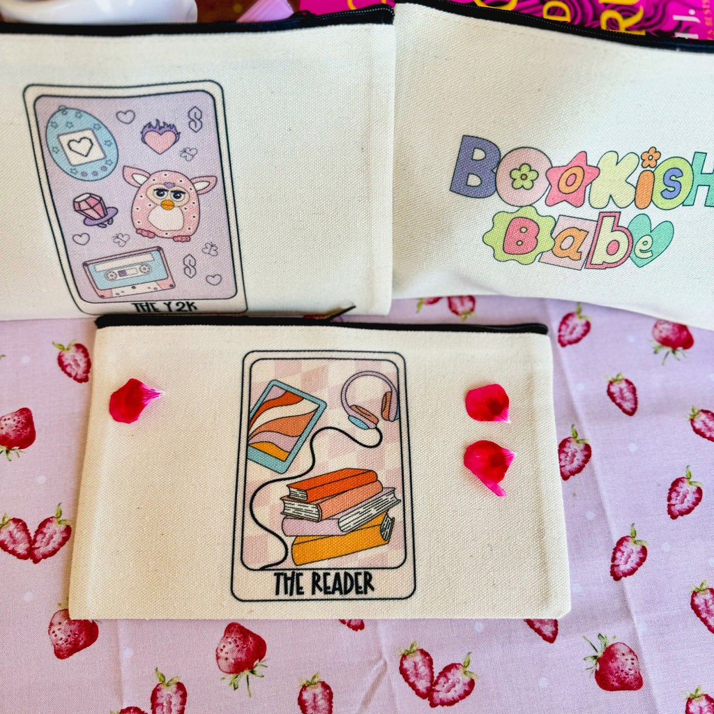Three canvas pouches with reading-related designs, placed on a surface with a strawberry pattern. Each pouch features a different design, including a cartoon of a character with glasses, text 'Bookish Babe', and a stack of books with the text 'THE READER'.