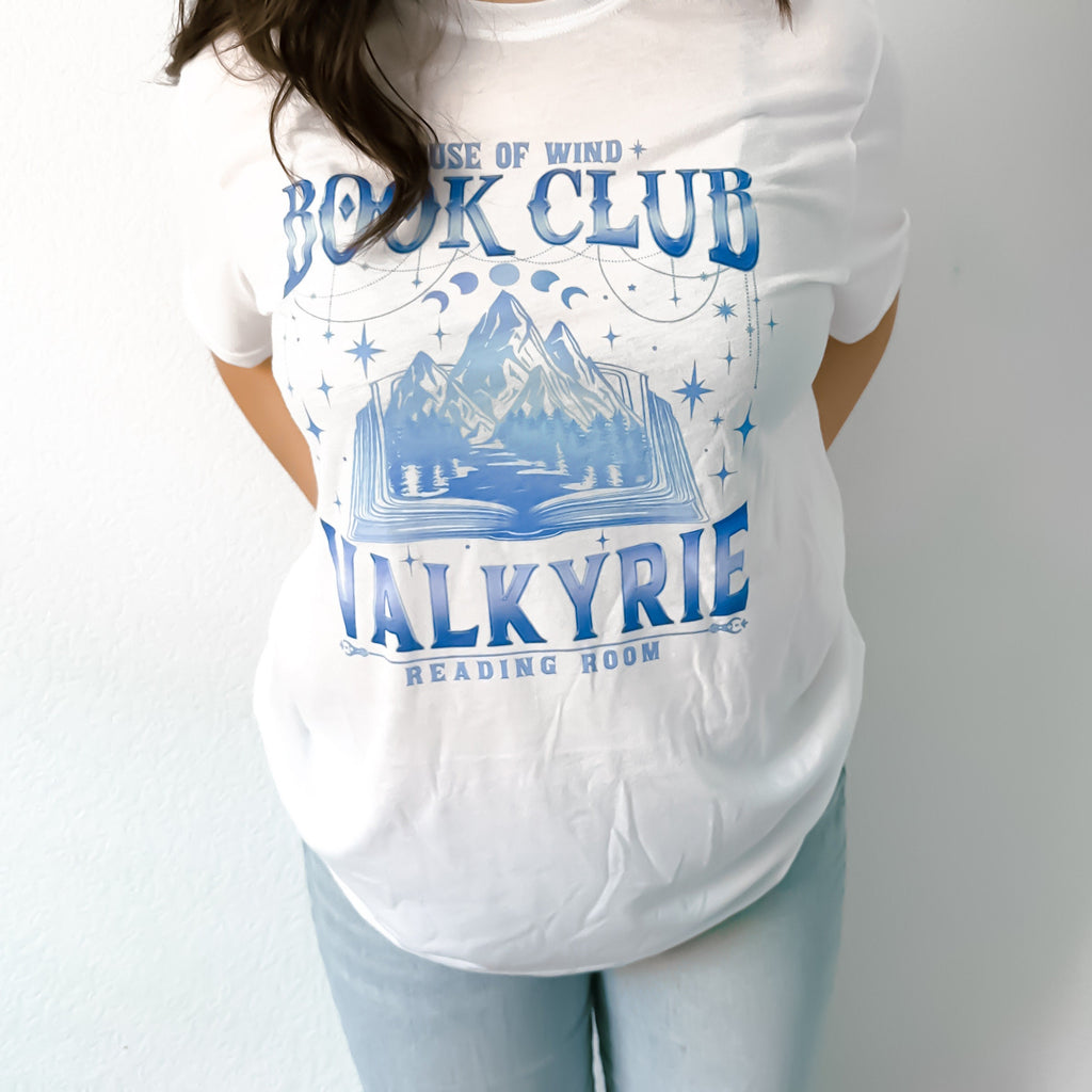 A person wearing a white graphic tee with 'House of Wind BOOK CLUB VALKYRIE READING ROOM' printed in blue.