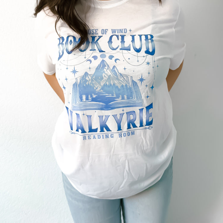 A person wearing a white graphic tee with 'House of Wind BOOK CLUB VALKYRIE READING ROOM' printed in blue.
