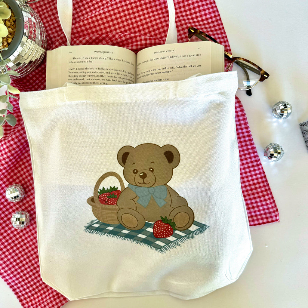A white canvas tote bag with a teddy bear holding a basket of strawberries, displayed on a red and white checkered surface with a book and decorative items in the background.