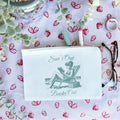 A white canvas pouch with a printed text 'Sun's Out Books Out' and an illustration of a person sitting on a chair. The pouch is placed on a surface with a floral pattern, and there are glasses and a pen beside it.