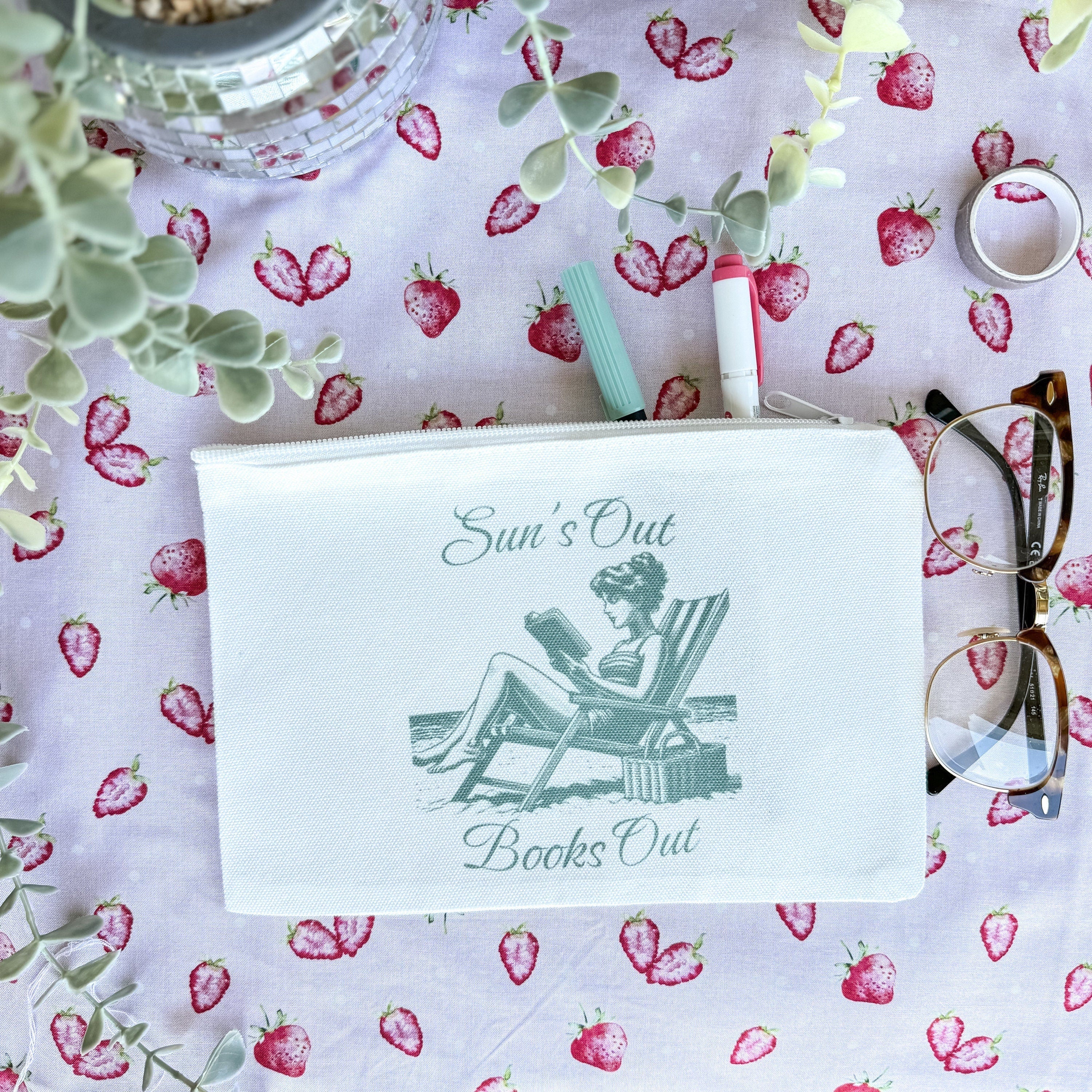 A white canvas pouch with a printed text 'Sun's Out Books Out' and an illustration of a person sitting on a chair. The pouch is placed on a surface with a floral pattern, and there are glasses and a pen beside it.