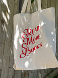 A beige canvas tote bag with 'READ MORE BOOKS' printed in red typography on the side, displayed against a wooden background.