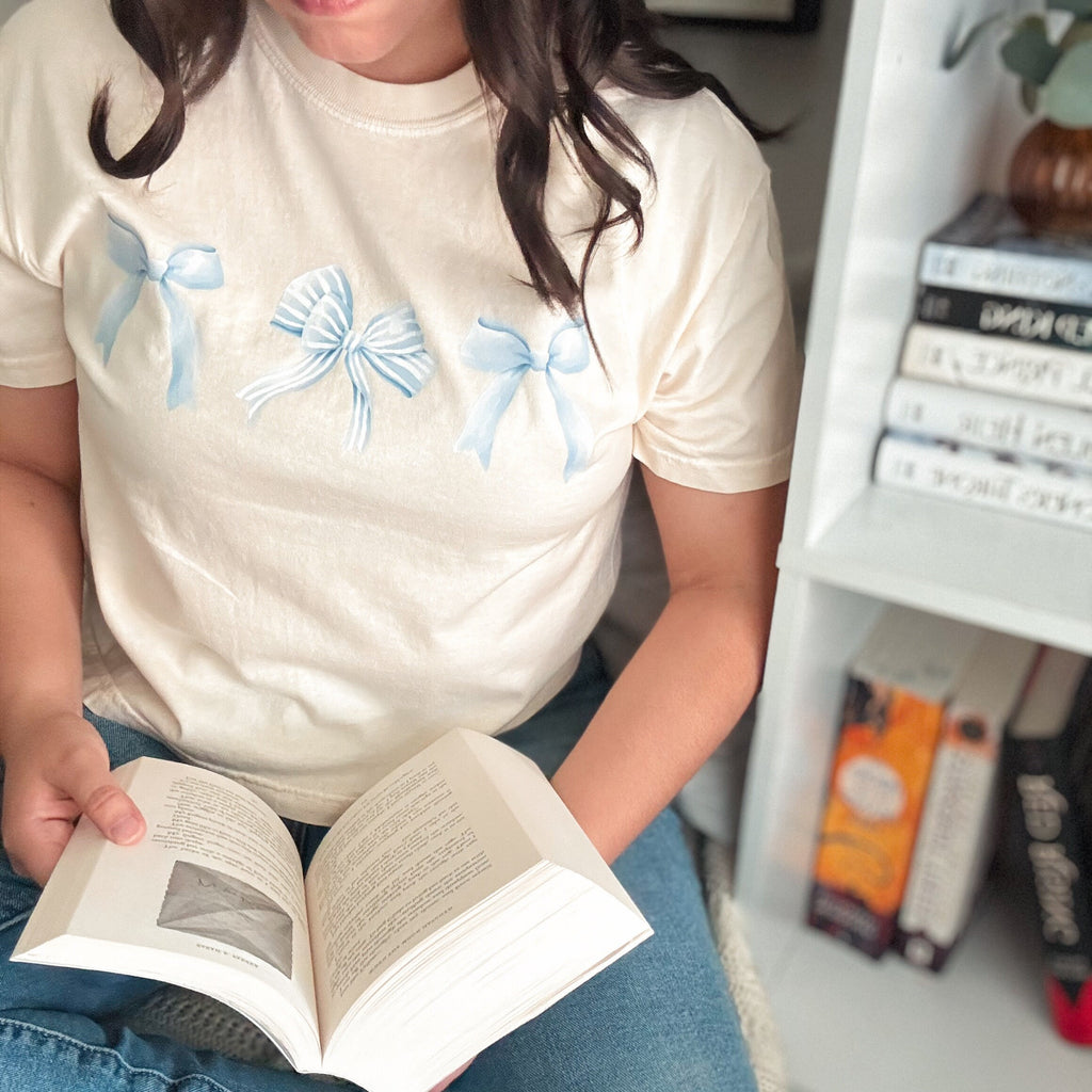 A person wearing a beige cropped t-shirt with blue bow graphics, sitting in front of a bookshelf and reading a book.