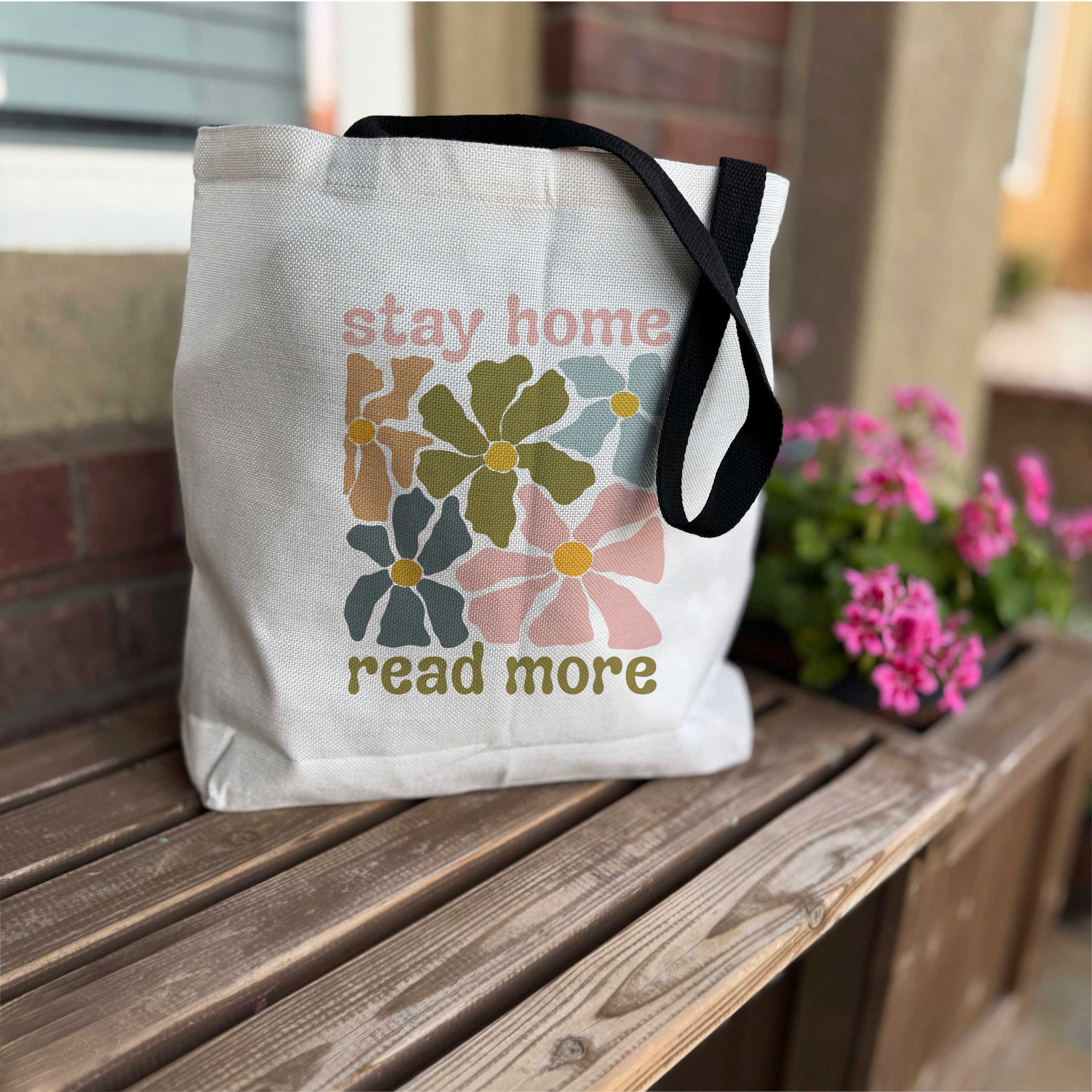 A white cotton tote bag with vibrant retro floral print design, featuring the text 'stay home read more' on it, placed on a wooden surface with flowers in the background.