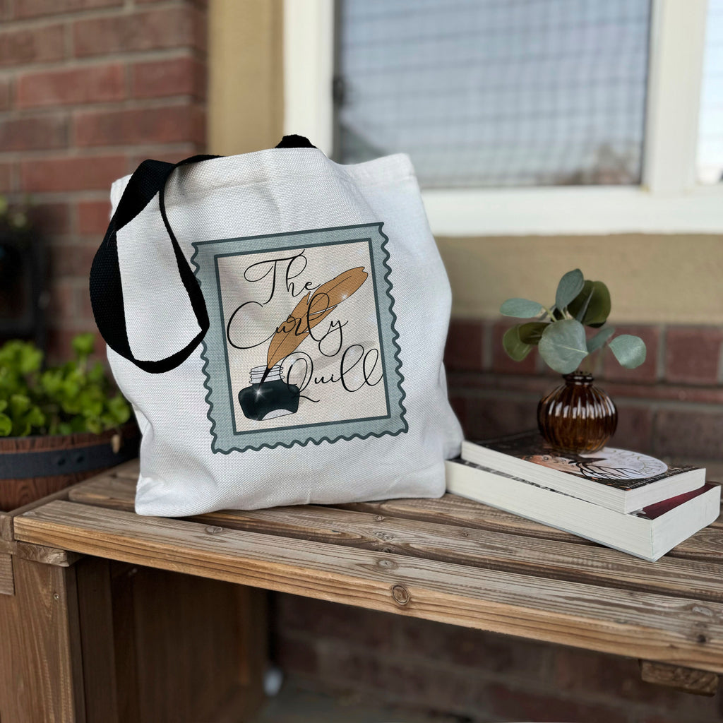 A white cotton tote bag with black straps featuring a printed design that includes a stylized representation of a book and a quote, placed on a wooden surface with a small plant and a book in the background.