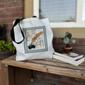 A white cotton tote bag with black straps featuring a printed design that includes a stylized representation of a book and a quote, placed on a wooden surface with a small plant and a book in the background.