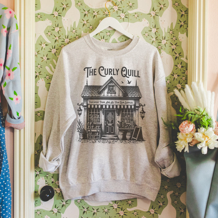 A gray sweatshirt with a vintage-style graphic print of a house and the text 'The Curly Quill' displayed on a hanger against a wall with floral wallpaper.