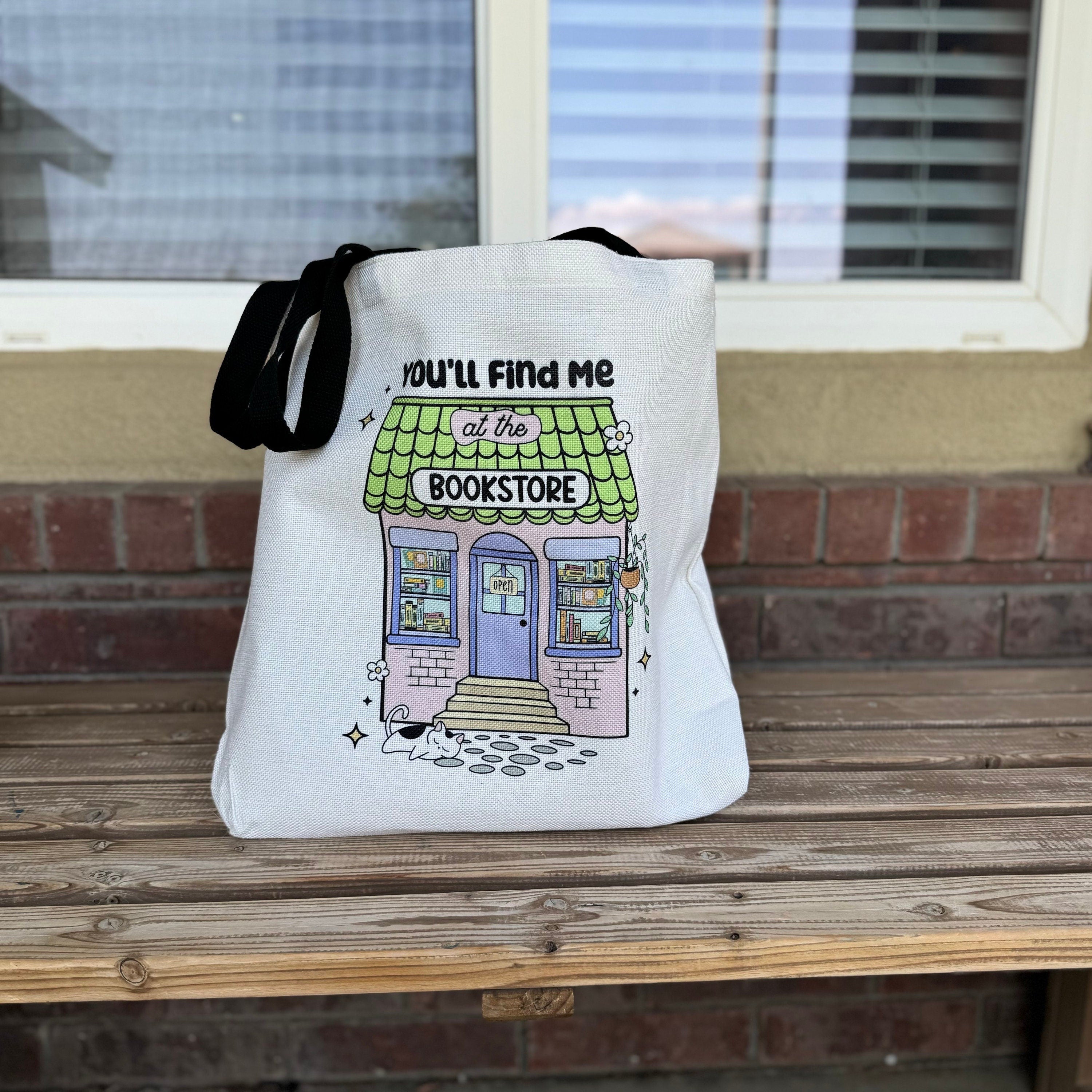 A white cotton tote bag with black text and a colorful illustration of a bookstore with a purple door, green awning, and white sign.