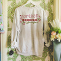A gray sweatshirt with the text 'forever BOOKISH' printed on the front, featuring a floral design, displayed on a hanger against a wall with floral wallpaper.